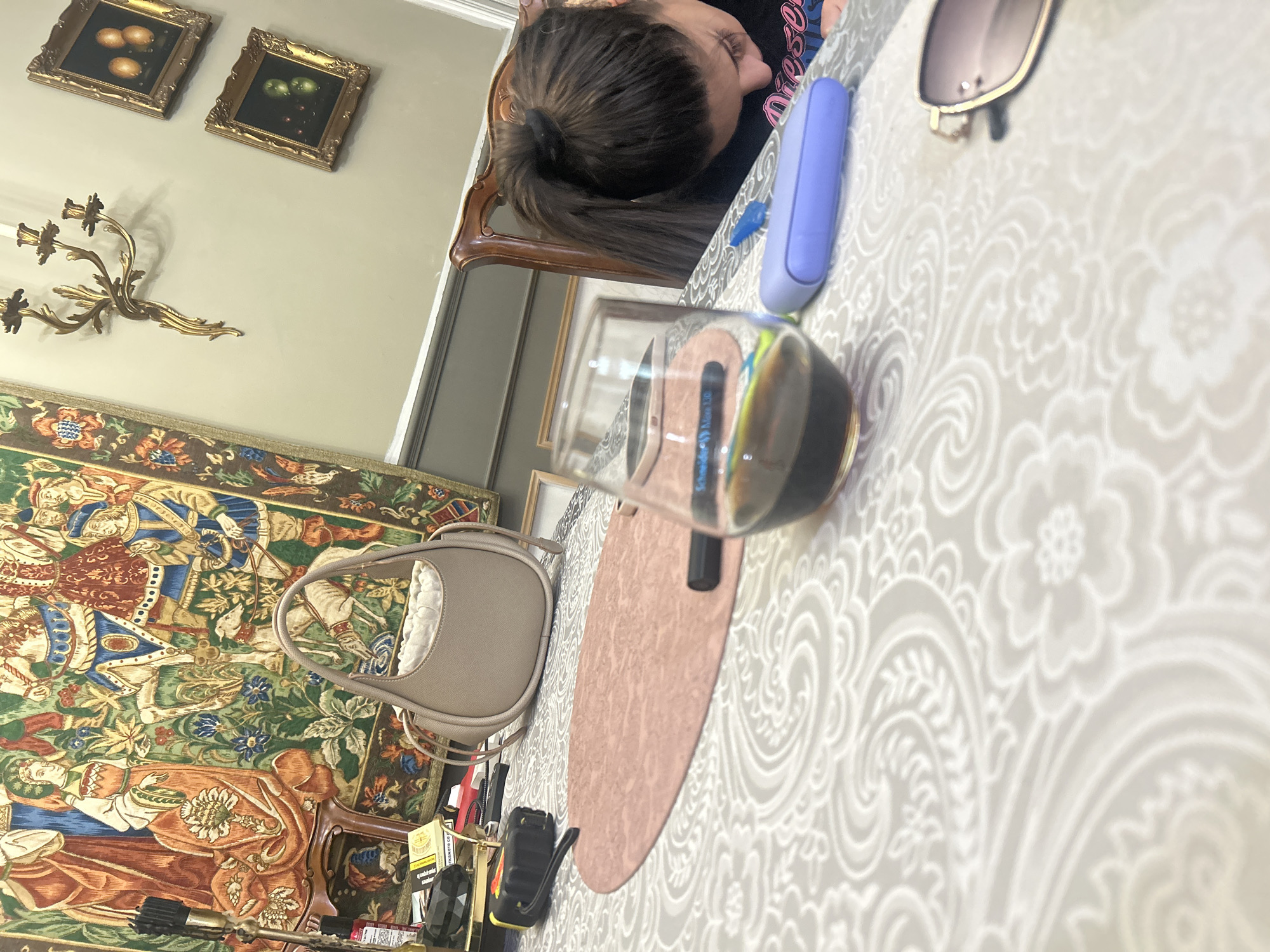This image depicts an indoor scene, likely a private residence. The specific location within Plovdiv, Bulgaria, cannot be determined from visual information present in the image.
In the foreground, a light grey or beige tablecloth with a white paisley and floral pattern covers a table. On the table, from right to left, a pair of gold-framed sunglasses with dark lenses is partially visible. A small, rounded, light blue/purple electronic device rests near the right edge. Centrally positioned, a clear glass contains a dark liquid, possibly red wine, with a colorful object reflecting or lying behind it. Beneath the glass is an oval-shaped, textured, light pink placemat, upon which a black marker or pen is placed horizontally. Further left on the table, a light beige or taupe-colored, crescent-shaped handbag with a drawstring top closure is visible. To the left of the bag, various items are clustered, including a black and orange tape measure or utility tool, a red and white spray can (possibly Schwarzkopf branded), a yellow and white cigarette pack (labeled "Виктори"), and a black geometric object.
One individual, appearing to be female with long dark hair tied in a high ponytail, is seated at the table on the right side of the frame. Her head is bent downwards, and she is focused on an activity on the table, the nature of which is obscured. She wears a dark shirt with partial text visible, possibly "Diesel". No other individuals are present.
The background features a large, ornate wall tapestry spanning the central and left areas, depicting a vibrant medieval or Renaissance scene with multiple figures, animals (dogs), and abundant foliage in green, orange, blue, and red tones. To the right of the tapestry, on a light-colored wall, an antique-style, three-armed, gold-colored wall sconce is mounted. Below the sconce, a dark wooden chair with carved details is partially visible. Further right, two smaller, dark-framed oil paintings are hung vertically on the wall; the upper painting depicts three orange fruits, and the lower one depicts three green fruits, both against dark backgrounds. A dark grey wainscoting runs along the lower wall section. The room is illuminated by ambient lighting, casting soft shadows.