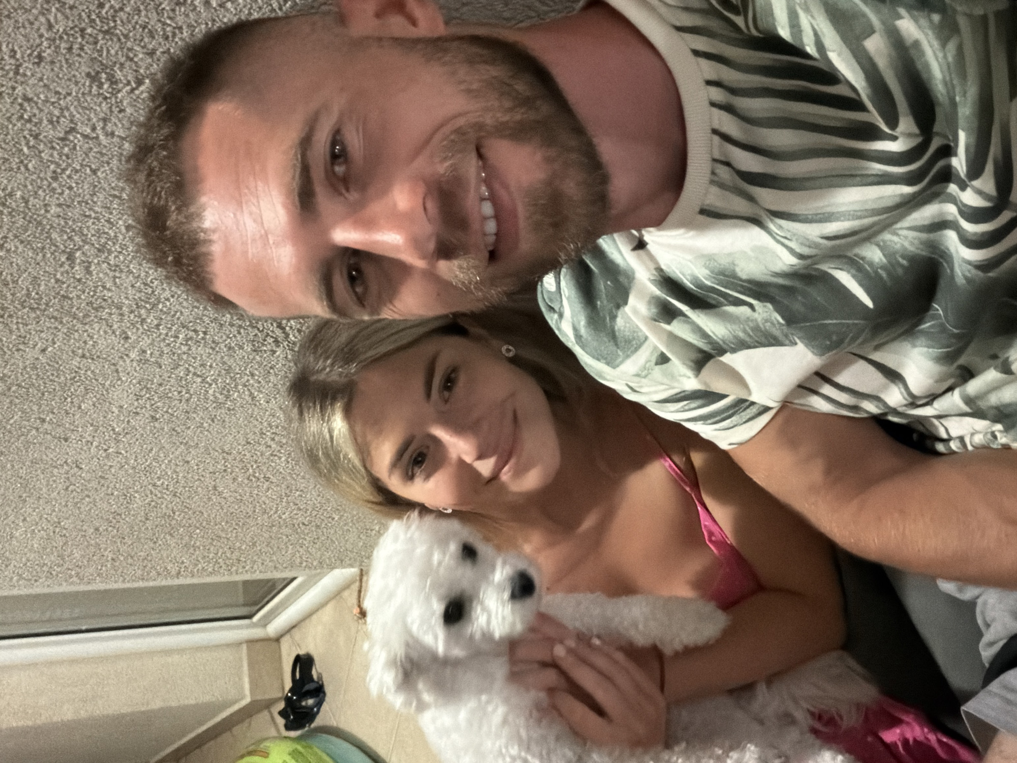 A man and a woman are posing for a selfie with a small white dog. The man is in the foreground on the right, smiling and showing his teeth. The woman is to his left, holding the dog. She has blonde hair and is wearing a pink bikini top. The dog is fluffy and white. The background appears to be an outdoor wall. Based on the context of a previous image that showed a view of Budva from a balcony with a similar wall texture, it is likely this photo was taken at a residence in Budva, Montenegro.