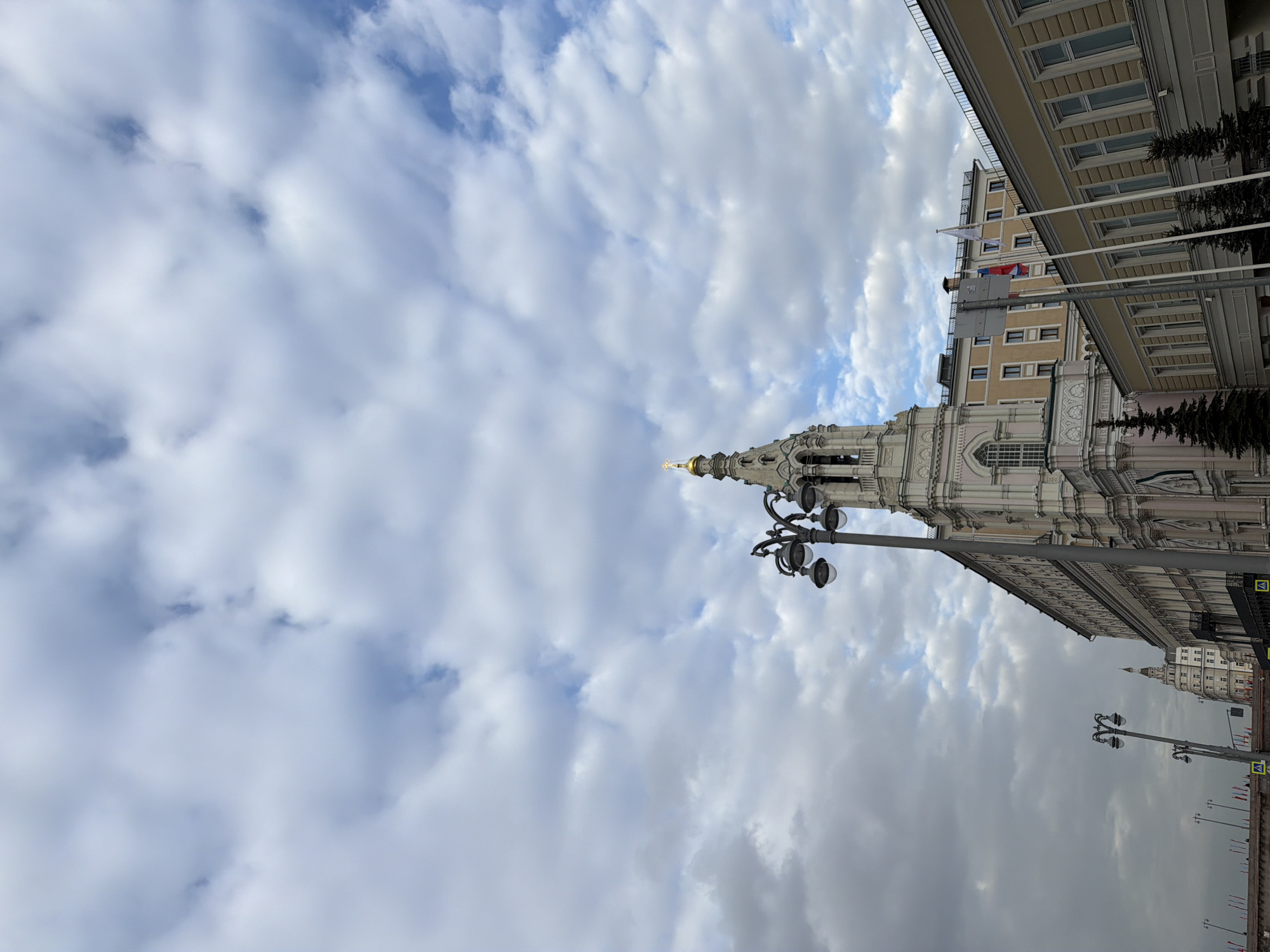 The image captures a view in an urban setting, showcasing a church with intricate architectural details, potentially located in the city of Unknown. The structure has a pointed spire topped with a golden dome, positioned against a backdrop of a sky filled with clouds, indicating varied weather conditions. Adjacent to the church stands a tall building with multiple windows and a beige facade, adorned with flags. The scene includes street lights with multiple globe-shaped lamps along a street or pedestrian area.