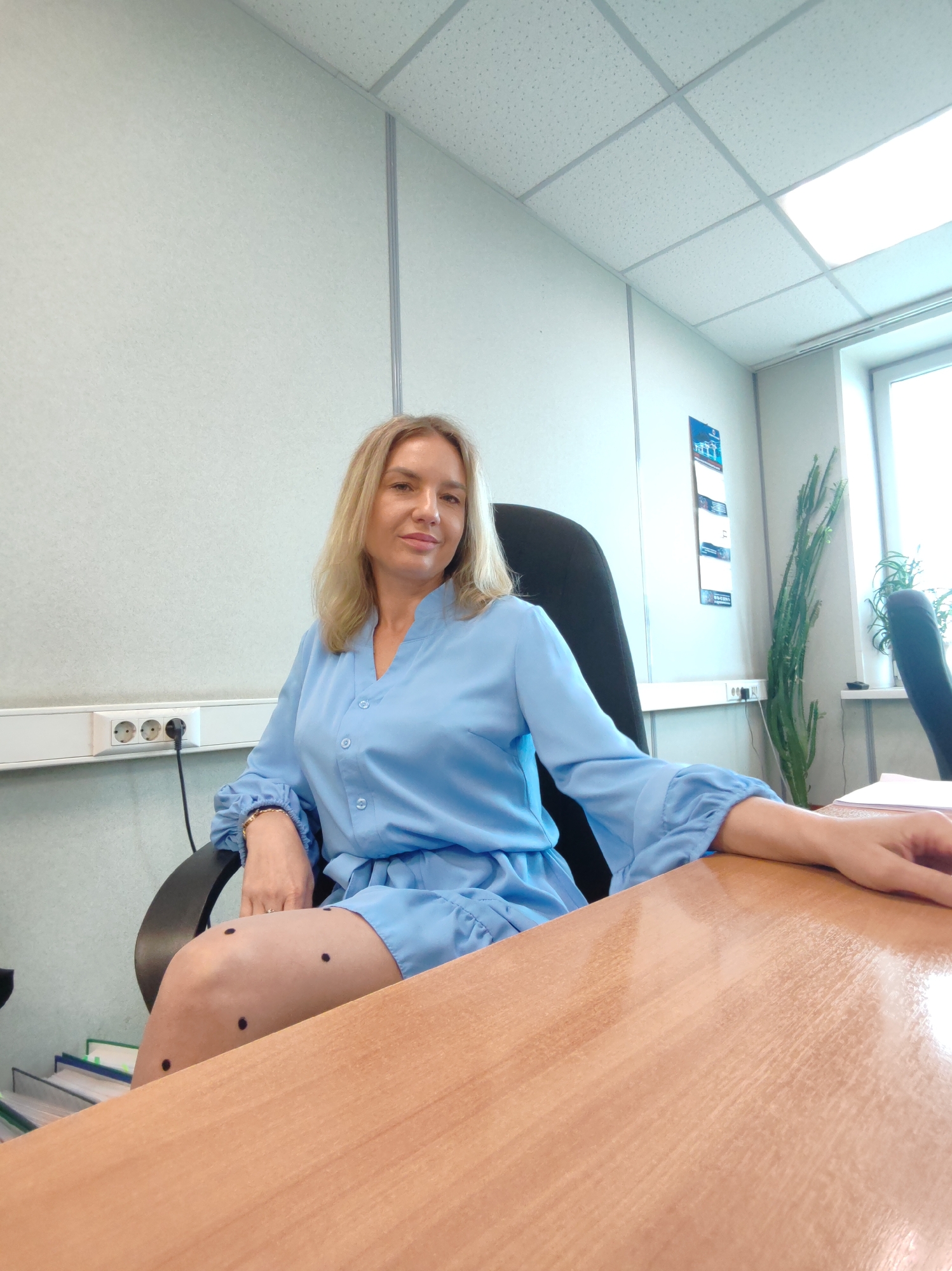 A woman with blonde hair is sitting at a desk in an office. She is wearing a light blue dress and has black polka dots on her legs. Her arm is resting on the desk. The room appears to be an office with a window, a ceiling with lights, and electrical outlets on the wall. There are also some plants in the background. The woman is looking towards the camera and smiling. The time of day is not discernible.