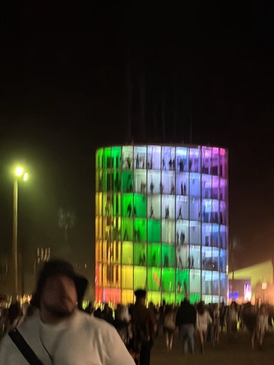 At night in Indio, United States, a large crowd has gathered around a tall, cylindrical structure illuminated with a rainbow of colors. The structure consists of multiple levels, each divided into smaller compartments where silhouettes of people can be seen. In the foreground, a person wearing a white shirt and a shoulder bag is partially visible, their face blurred. Various other figures are scattered throughout the crowd, some wearing hats. A lamppost shines brightly to the left of the illuminated structure. The overall scene is active and vibrant, indicative of an event or festival.