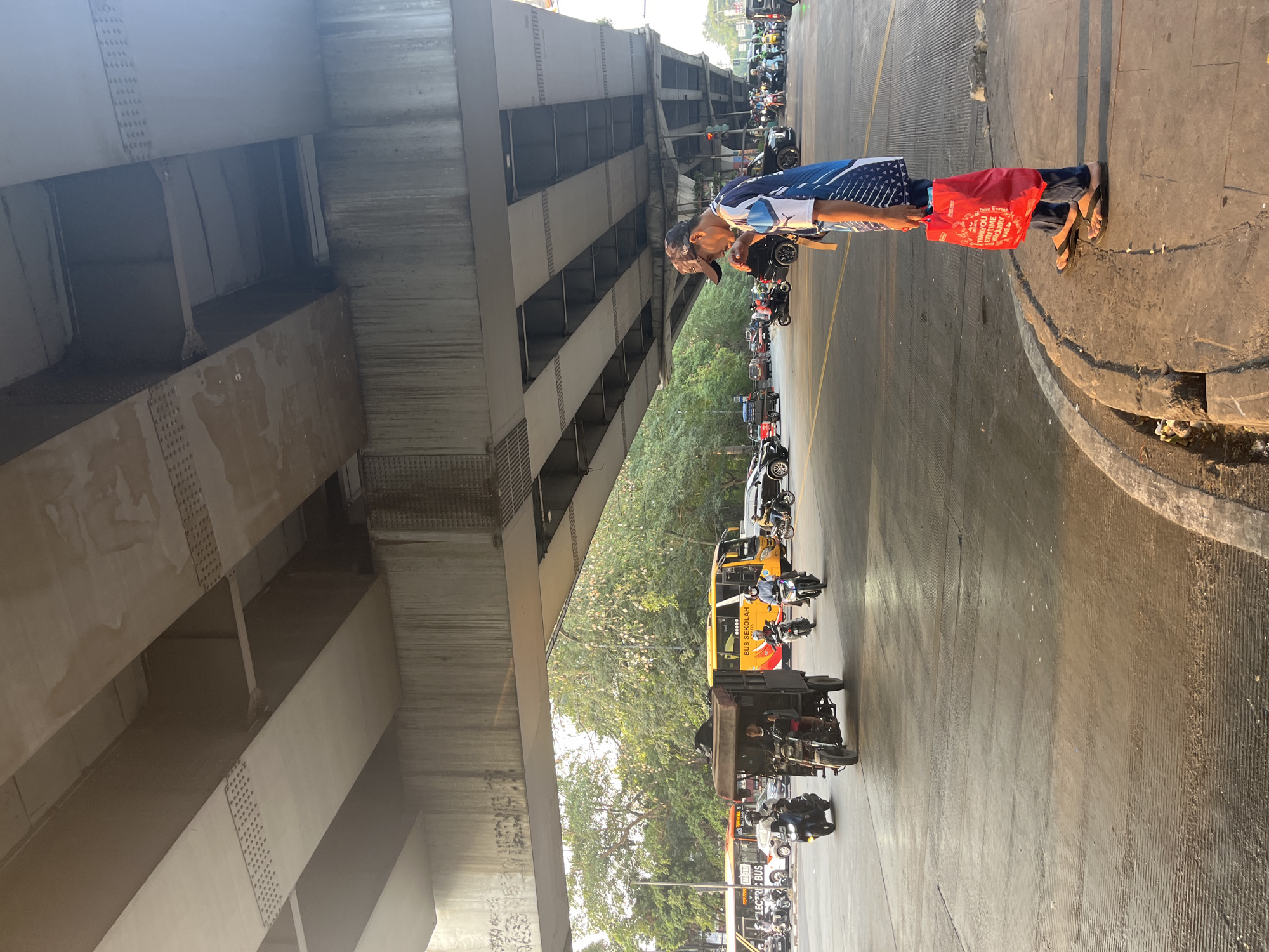 The image depicts a busy street scene in East Jakarta, Indonesia, under a large overpass. The overpass, constructed from concrete beams, casts a shadow over the road below. In the foreground, a man wearing a blue and white shirt, dark pants, and a cap stands on a sidewalk, holding a red plastic bag. He is facing away from the camera, looking towards the street.
The street is filled with various vehicles, including motorcycles, cars, and a yellow school bus. Several motorcycles are seen navigating the intersection. A three-wheeled motorized vehicle, often used for transport in Indonesia, is also prominent. Trees and foliage line the background, indicating an urban environment. The lighting suggests it is late afternoon, with the sun casting long shadows. The overall scene conveys the hustle and bustle of daily life in a dense urban area.