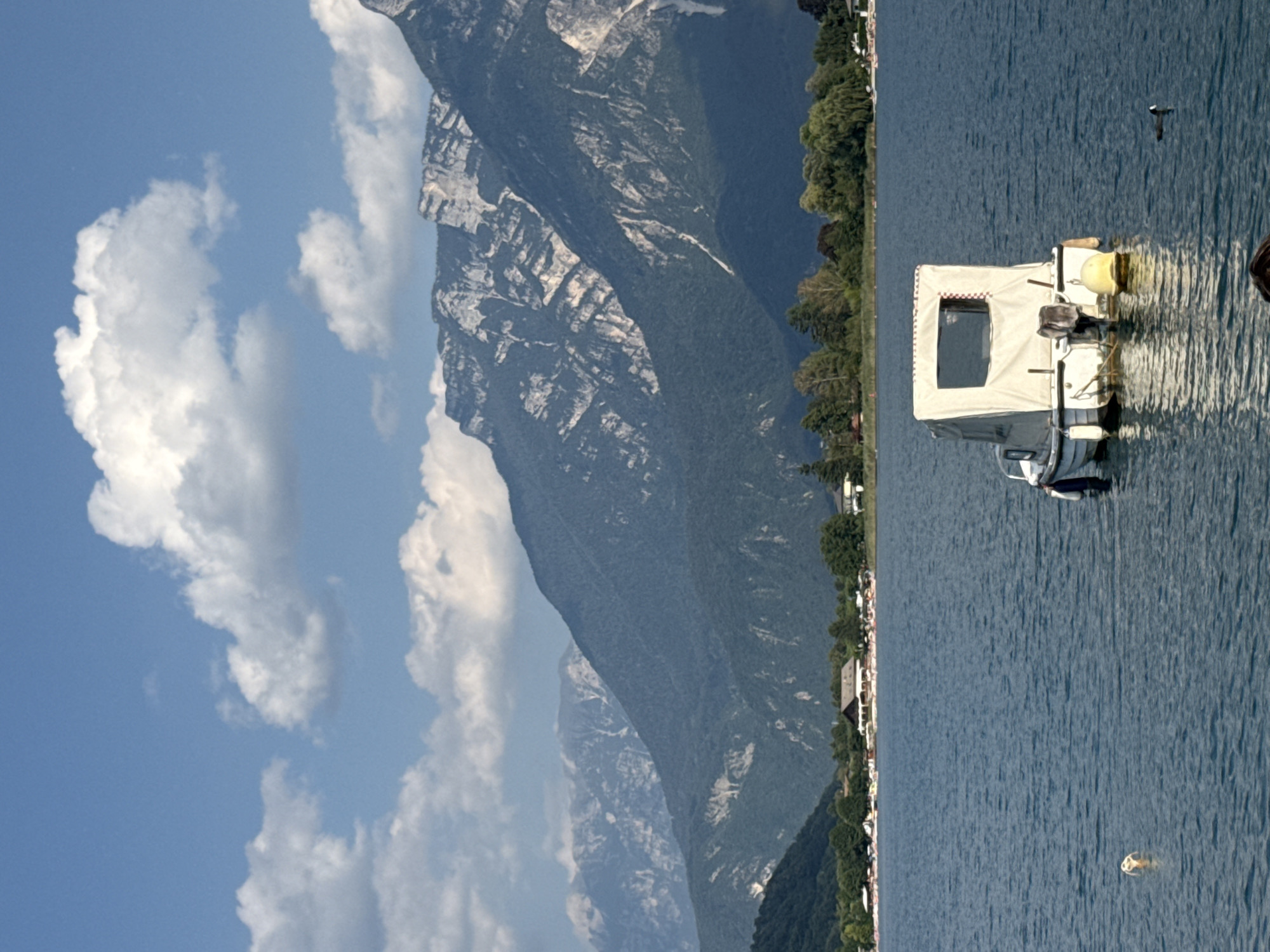 The image depicts a scenic view of Calceranica al Lago, Italy, on a clear day with a blue sky dotted with white, puffy clouds. In the foreground, a calm, blue lake occupies the majority of the frame, reflecting the light. A small, white motorboat with a beige canopy is moored in the middle of the lake, facing away from the camera. The boat has a visible outboard motor and a yellow buoy is attached to its stern. To the left of the boat, a small, round, yellow object floats on the water. In the distance, the shoreline is lined with trees and buildings, including what appears to be a resort or residential area. Behind the shoreline, a range of majestic, dark green mountains rises, their rugged peaks illuminated by the sun. The overall impression is one of a peaceful and picturesque summer day in a lakeside town.