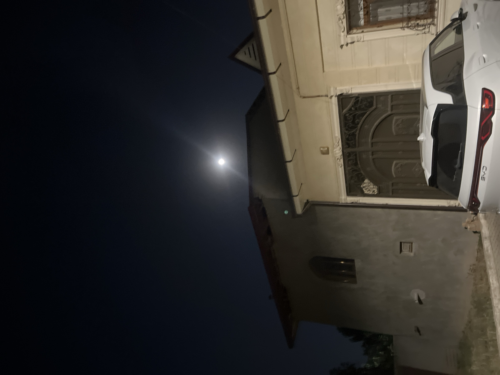 A white BYD SUV is parked in front of a large, ornate gate at night. The moon is visible in the dark sky, casting a soft light on the scene. The SUV has its rear lights on and appears to be in good condition. The gate is made of dark metal and has intricate scrollwork. A house with a light-colored facade is visible behind the gate, with a window and decorative molding. The scene is located in an urban or suburban environment, with some trees visible in the background.