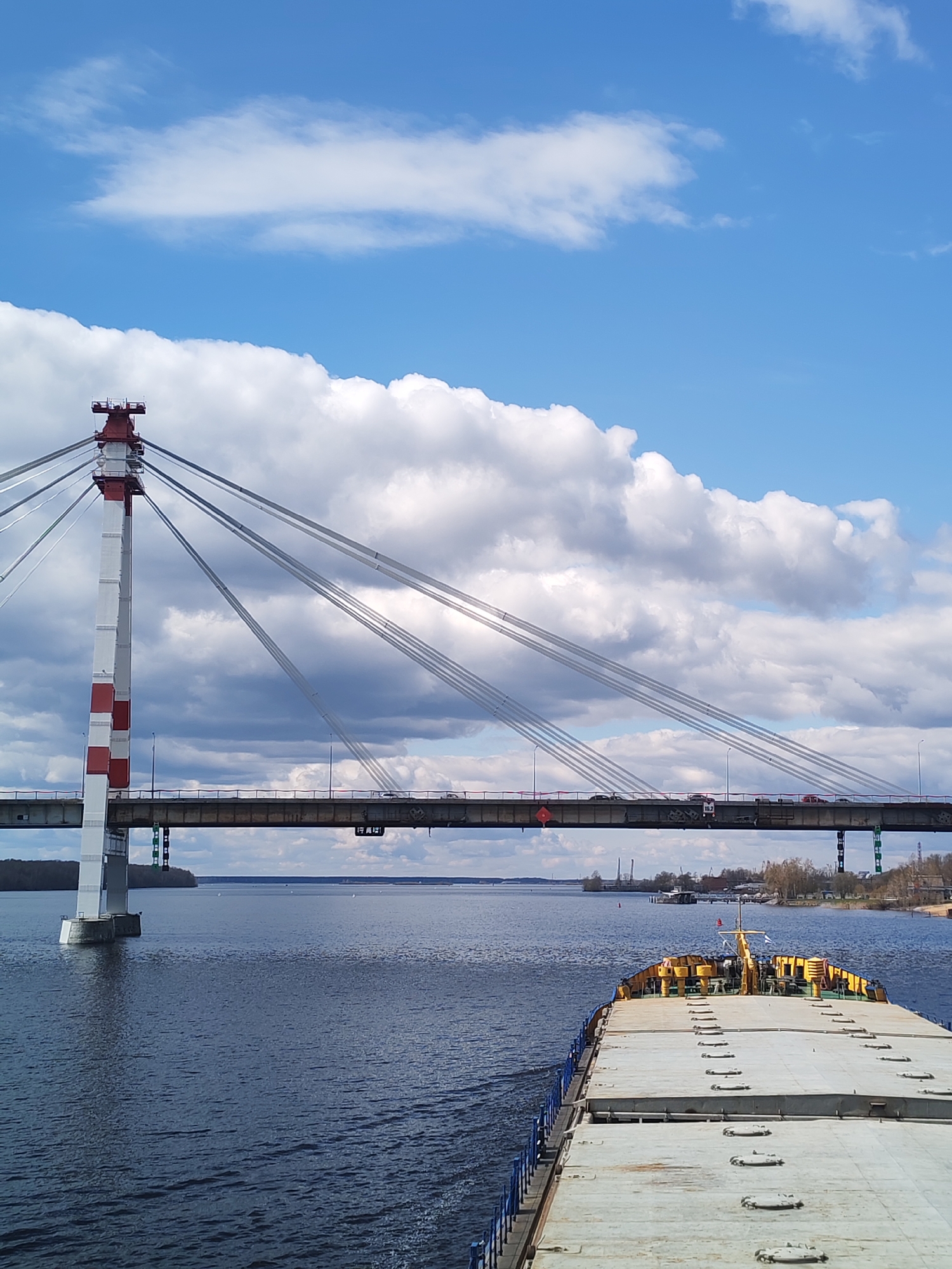 A daytime view of a cable-stayed bridge in Cherepovets, Russia, spans a wide body of water. The bridge is supported by a single, tall white pylon with red accents at the top and middle. Cables extend from the pylon to the bridge deck. Cars are visible crossing the bridge. Large, puffy clouds fill the blue sky above the bridge. In the foreground, the deck of a barge is seen, indicating the photo was taken from a boat on the river. The water is dark, with light ripples. The riverbanks have some vegetation.