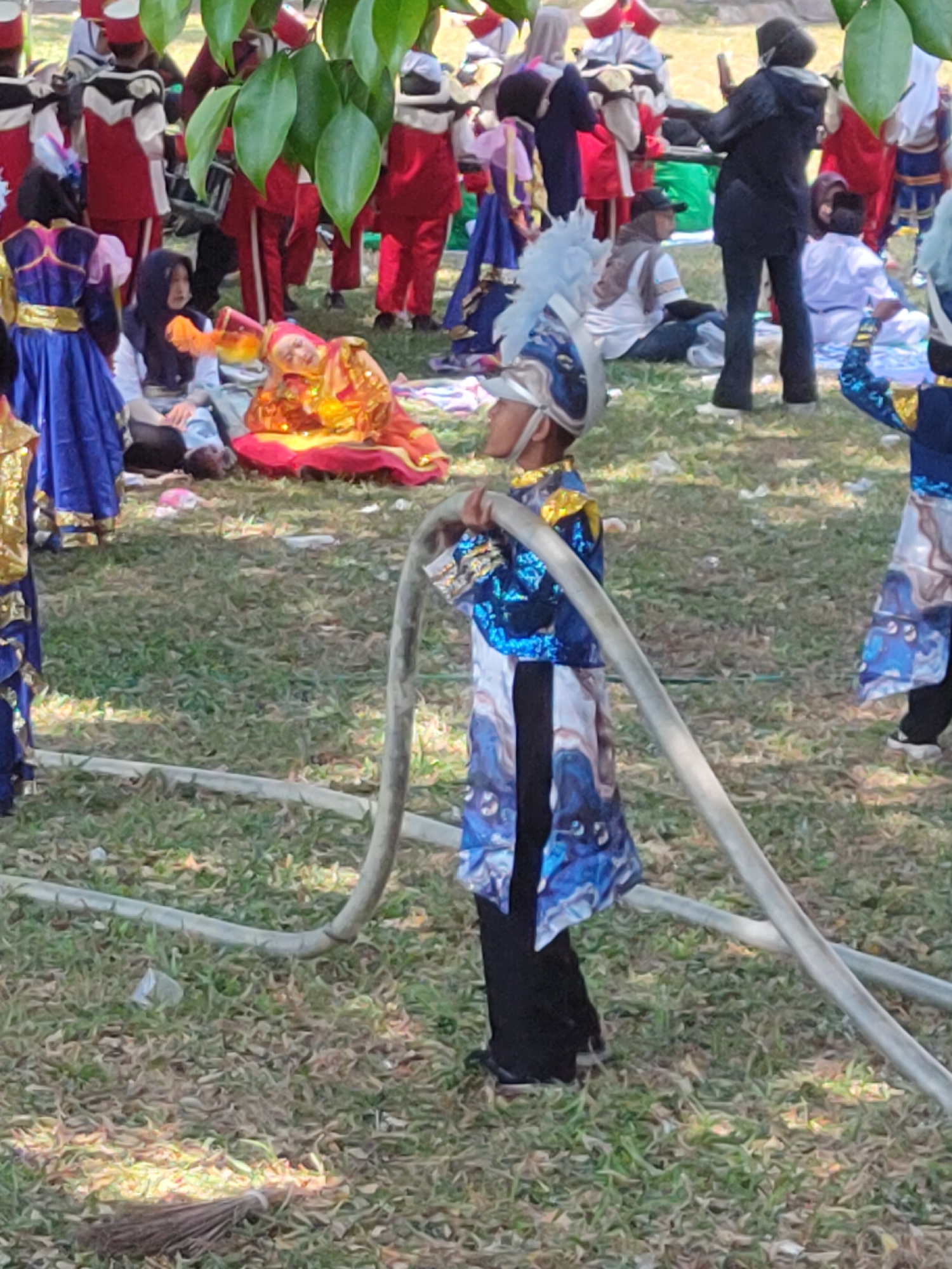The image depicts a street performance or parade in Ngasem, Indonesia, likely a public gathering in a grassy area with some trees. A group of children in colorful marching band uniforms are present. In the foreground, a child in a blue and silver sequined uniform with a feathered headpiece is holding a large, coiled hose. To the left, another child in a similar blue and gold uniform is standing, and further back, a child in a bright orange and red costume is lying on the grass. Other children and adults in various attire, some in red and white uniforms, are visible in the background, suggesting a larger event is taking place. The ground is covered in dry grass, with fallen leaves and some scattered litter. The overall impression is a lively, outdoor event involving a performance troupe.