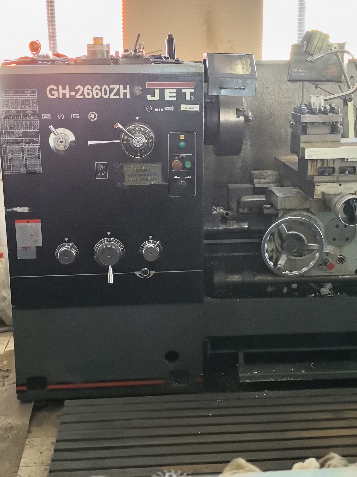 The image shows a JET GH-2660ZH lathe in what appears to be a workshop. The lathe is a dark gray industrial machine with various dials, levers, and buttons on its control panel. There are also some labels with Russian text, including one that reads "КНОПКА АВАРИЙНОГО ОТКЛЮЧЕНИЯ," which translates to "EMERGENCY SHUTDOWN BUTTON." To the right of the lathe, a part of another machine, possibly a milling machine, is visible, with a tool holder containing cutting tools. The background shows a window with blinds and a wall. The floor is concrete and appears somewhat dirty, with some debris. The overall environment suggests an active industrial or workshop setting.