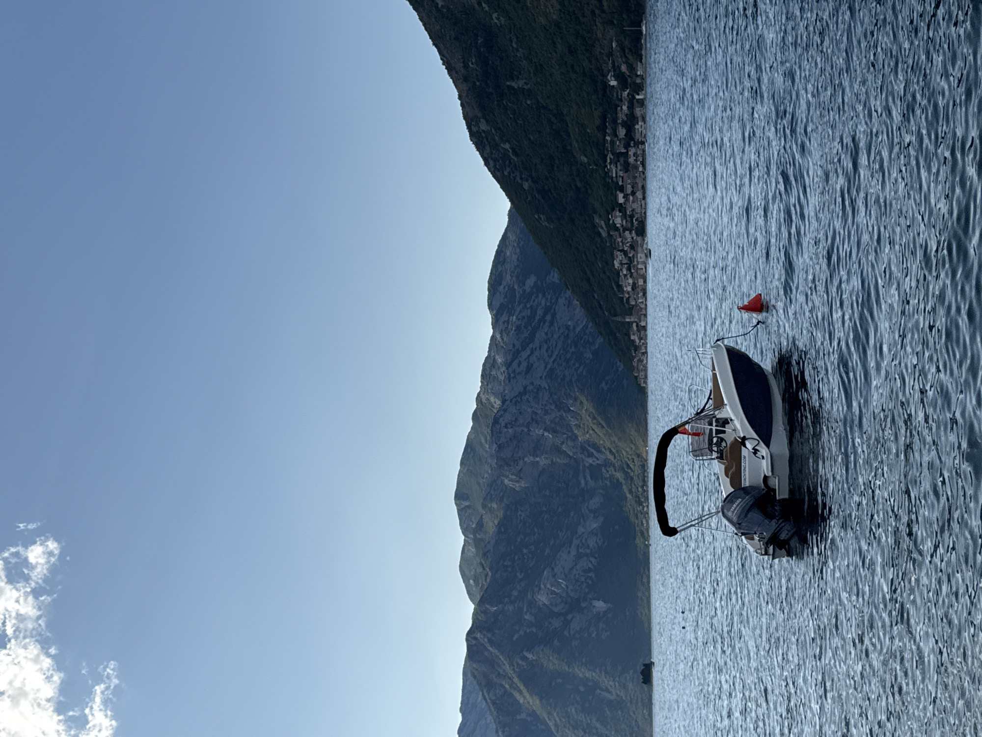 A small white and blue motorboat with a black canopy is anchored on calm blue water in front of the town of Prčanj, Montenegro. The town is situated at the base of steep, verdant mountains that rise sharply behind it. The sky above is clear and bright blue with a single white cloud in the upper left corner. A bright orange buoy floats near the boat. In the distance, on the left side of the frame, a small island with a single structure is visible. The water exhibits a rippled texture, indicating a slight breeze. The boat has a Yamaha outboard motor. The scene suggests a peaceful, scenic day in a coastal town.