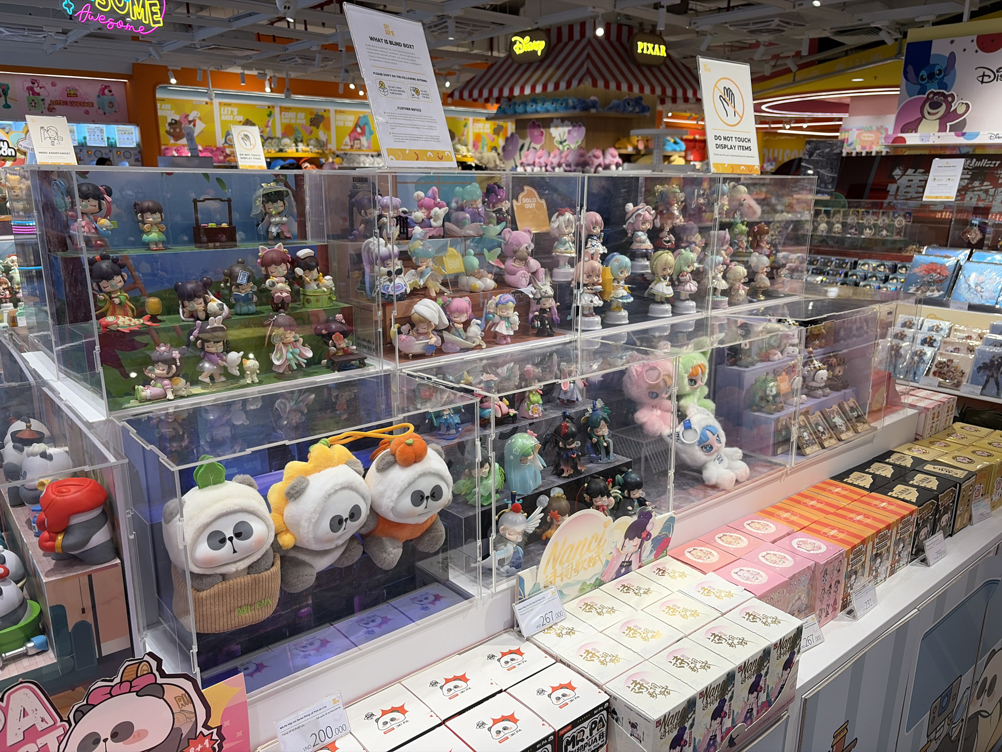 The image shows a retail display of collectible figures and blind box toys inside a store, likely in a mall. The toys are organized in clear plastic cases with multiple tiers, showcasing various characters and themes, including pandas and Japanese-style figures. Below the displays are boxes of blind box toys available for purchase. Price tags in Vietnamese Dong (VND) are visible on some of the boxes. Signs are displayed advising customers "Do Not Touch Display Items" and explaining "What is Blind Box." Brands such as Disney and Pixar are visible in the background.