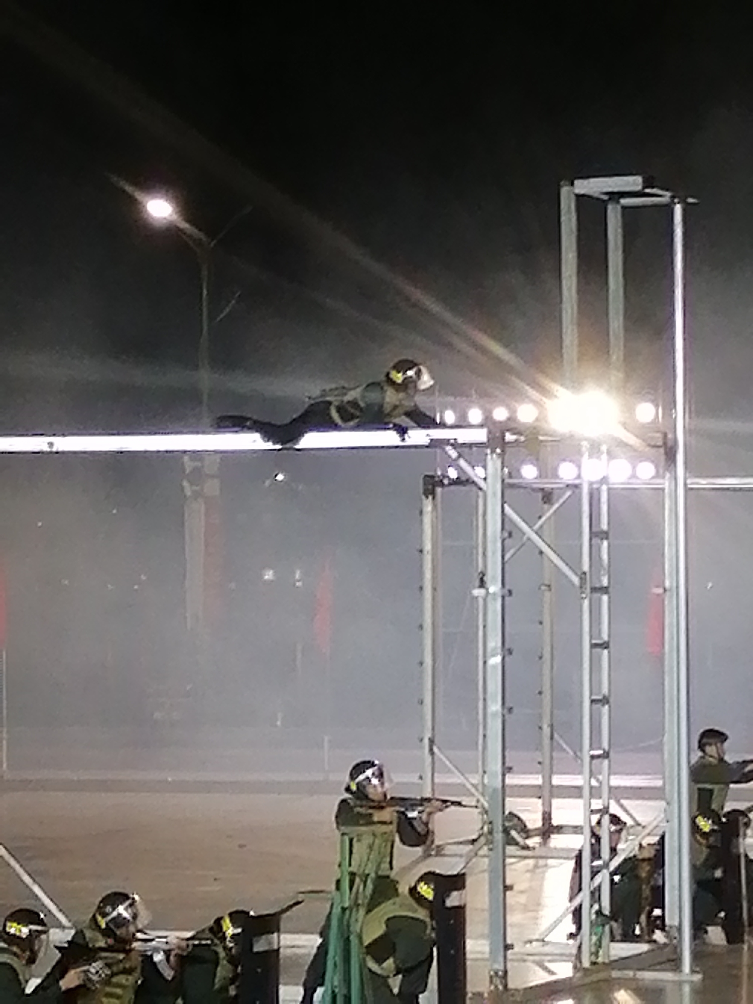 A person in tactical gear and a helmet is captured mid-air, appearing to be in the process of jumping or falling from a raised platform. They are wearing a dark outfit with what looks like a green vest and dark pants. The person's arms and legs are extended.
The scene appears to be an outdoor event at night, as indicated by the dark sky and bright artificial lights. A street lamp is visible in the upper left of the frame, emitting a strong glow. In the foreground, several other individuals also in tactical gear and helmets are positioned, some holding what seem to be rifles or similar equipment. They are standing behind barricades or shields.
A tall metal structure, possibly part of a stage or a training apparatus, is prominent in the right side of the image, with bright lights illuminating the area around the airborne subject. The ground is obscured by a smoky or foggy atmosphere, creating a hazy effect. There are hints of red objects in the background, possibly flags or banners. The overall atmosphere suggests a demonstration, training exercise, or performance. No visible text is present.