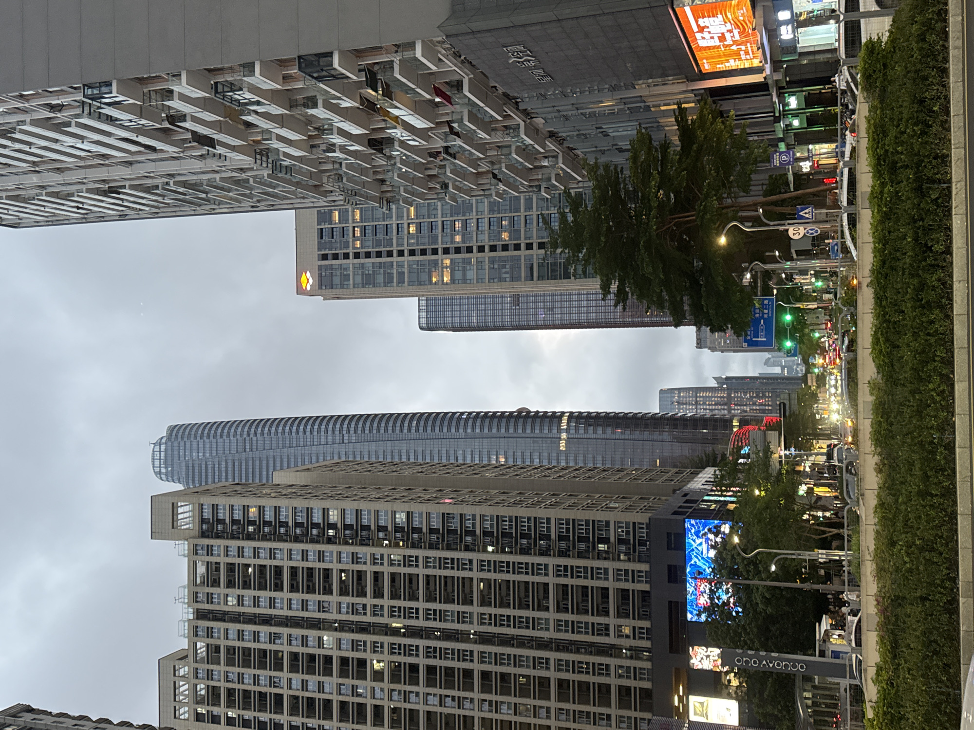 The image depicts a street scene in Futian District, China, characterized by modern architecture and a cloudy sky. The perspective is from street level, looking upwards at several high-rise buildings of varying designs.
On the left side, the upper building shows a pattern of multiple units with balconies or protruding structures. Below that is a tall building in a neutral color with many windows.
On the right side, street lighting and signage are visible, along with green foliage suggesting a park or landscaped area. A large digital screen displays bright, abstract imagery. Street signs and traffic lights are also visible, indicating vehicular movement. The environment suggests an urban center with mixed residential and commercial properties.