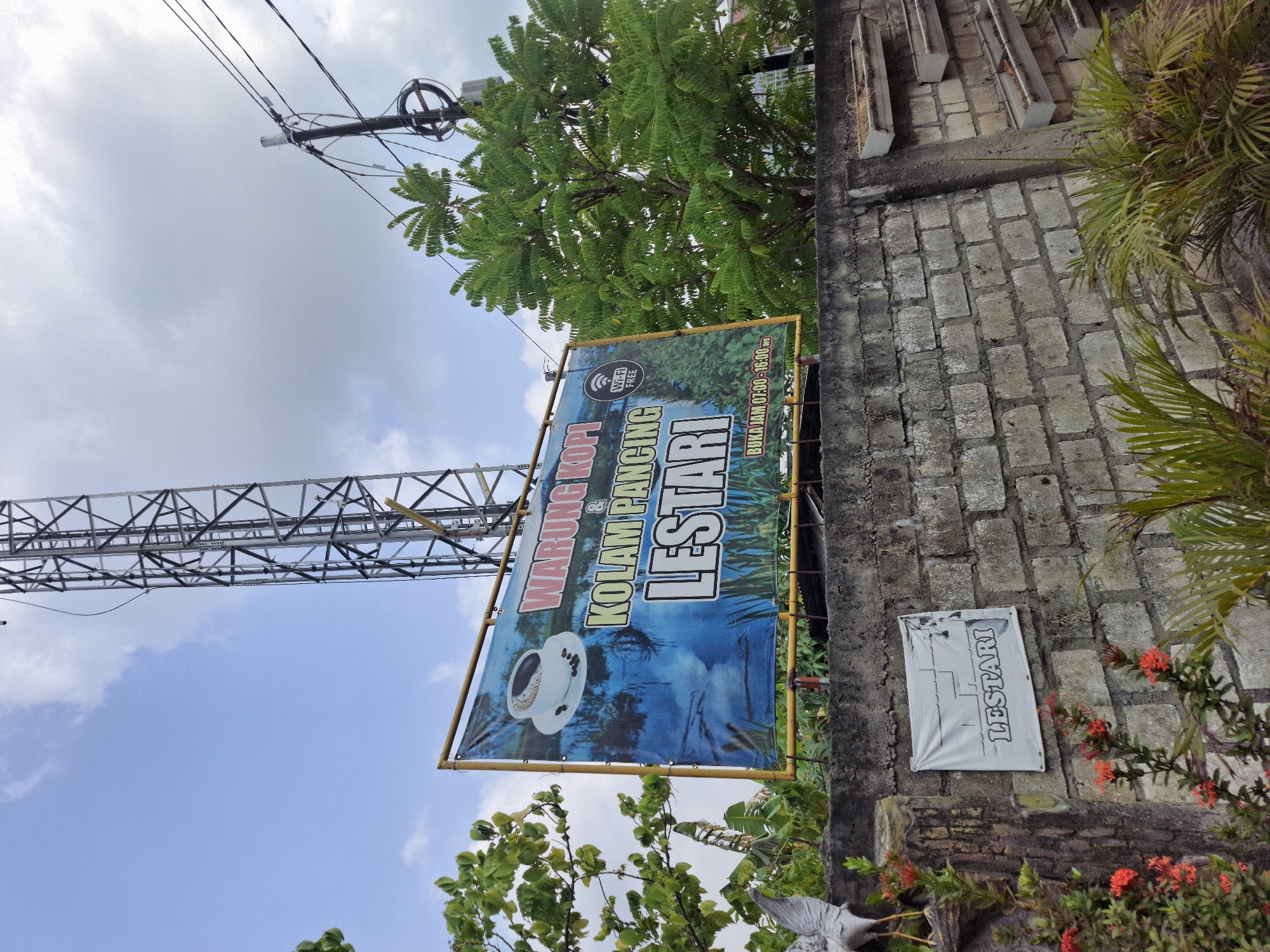 This image depicts an outdoor scene, likely in Lamongan, Indonesia. A large blue banner is prominently displayed, advertising "WARUNG KOPI & KOLAM PANCING LESTARI" (Coffee Shop and Fishing Pond Lestari). The banner features an image of a coffee cup and a body of water with reeds, and it also indicates "WI-FI FREE" and operating hours "BUKA JAM 07:00 - 16:00". The banner is supported by a yellow frame and is situated above a concrete brick wall. To the left of the banner, a tall metal tower, possibly a communication tower, is visible. Lush green trees frame the scene on the right and left. Below the banner, on the brick wall, a smaller white sign with the word "LESTARI" is attached. Red flowers and green foliage are present in the foreground, adding color to the scene. The sky is partly cloudy.