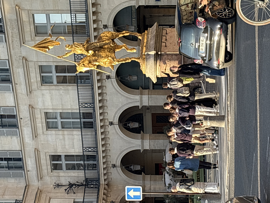 The image depicts an urban scene in Paris, France, specifically at the Place des Pyramides. The central feature is a large, gilded equestrian statue of Joan of Arc, mounted on a stone pedestal. Joan of Arc is depicted in armor, holding a banner aloft.
In the background stands a multi-story stone building, characteristic of Parisian architecture, featuring a ground floor with a series of arches and an upper floor with windows, grey shutters, and a decorative black wrought-iron balcony. Hanging lanterns are visible under the arches. A partial sign reading "STIEN GAUDARD" is visible on the building to the right of the statue.
In the mid-ground, a group of approximately 15-20 individuals is gathered. Most are young people, possibly students, dressed in dark jackets/sweaters and light-colored trousers, many carrying backpacks. They are positioned near the base of the statue, some looking towards it, others interacting within the group. Two adult females are on the left, one in a light coat and head covering, another in a blue denim jacket. An adult male in a dark jacket and jeans stands next to a parked car on the right. An adult female stands between the student group and the car, facing left with her mouth slightly open.
The foreground shows a paved roadway. A dark grey or black Smart car with a visible French license plate "GK-394-RL" is parked or stopped on the right side of the road. A portion of a bicycle wheel with a brown tire is visible in the lower right corner. A blue square sign with a white upward arrow is mounted on a pole in the upper left, indicating a one-way street. Concrete bollards separate the pedestrian area from the roadway. The scene is illuminated by strong sunlight, casting distinct shadows.