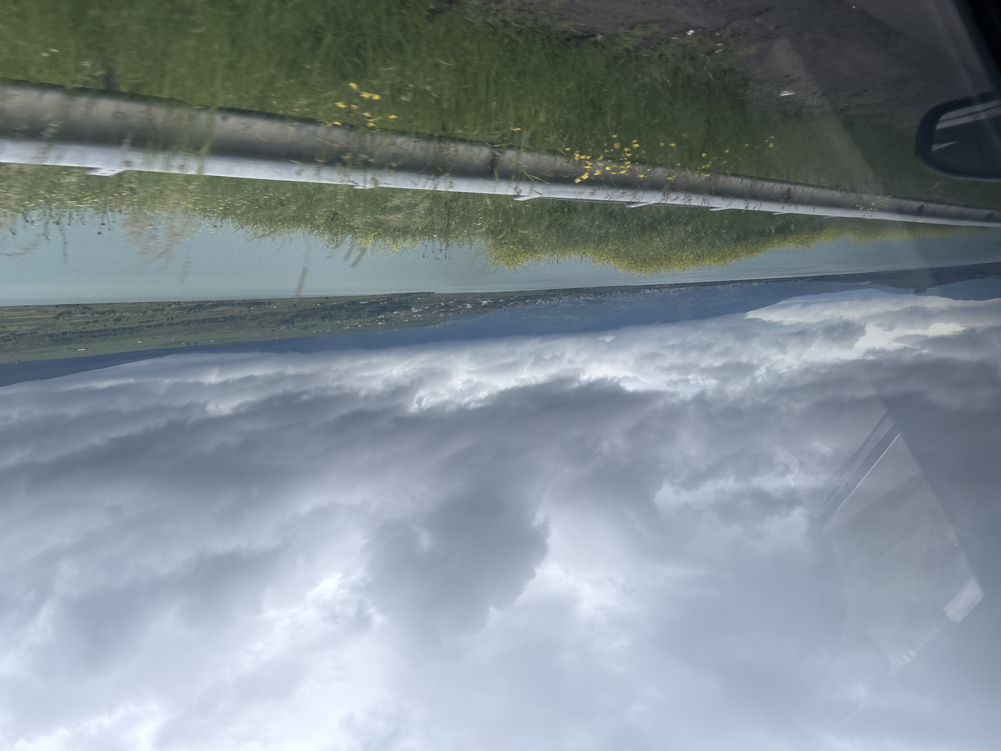 The image, taken from a moving vehicle, shows a landscape with a body of water in the midground, possibly a lake or reservoir. A grey guardrail with some green vegetation grows in the front and green grass is visible. A strip of land with buildings is seen across the water. Above, a thick, overcast sky filled with grey clouds dominates the upper part of the image. The vehicle's window frame is visible in the right side, indicating the perspective of the photo. No people are visible. The location, Tbilisi, Georgia is not visible.