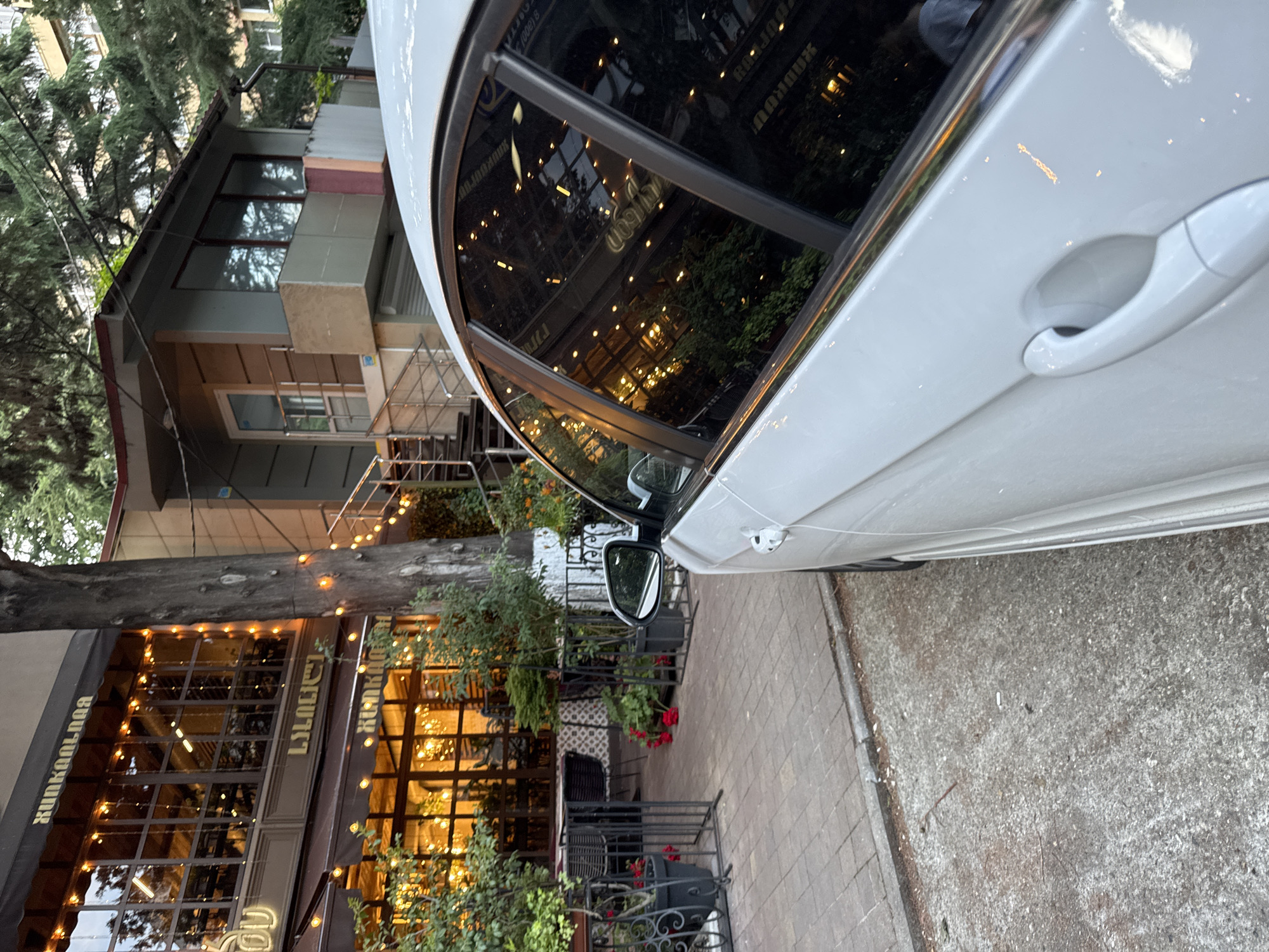 The image, rotated 90 degrees counter-clockwise from its original orientation, captures a street scene in Sochi, Russia, during what appears to be twilight or early evening. The foreground is dominated by the right side of a white car, likely a sedan, with its passenger-side door handle and a portion of its side mirror visible. The car's windows, including a large side window and possibly a sunroof, reflect the opposite building, showing an array of warm, glowing string lights and interior illumination.
Adjacent to the car is a paved sidewalk, which transitions into an unpaved, grayish ground area. A prominent, thick tree trunk extends horizontally across the mid-ground.
To the left, a warmly lit building, presumably a cafe or restaurant, features large windows showcasing its illuminated interior with numerous decorative string lights. Potted plants with red flowers adorn the exterior. Above the windows, an awning displays text that appears to be "KUNKANOGA" and "LILILELI," possibly in Georgian script, indicating the establishment's name.
Further in the background, partially obscured by lush green trees, is a multi-story residential building. The overall lighting, characterized by the bright artificial lights of the cafe against a darkening sky, strongly suggests either dusk or early night. The scene is static, devoid of human activity, presenting a peaceful urban snapshot.