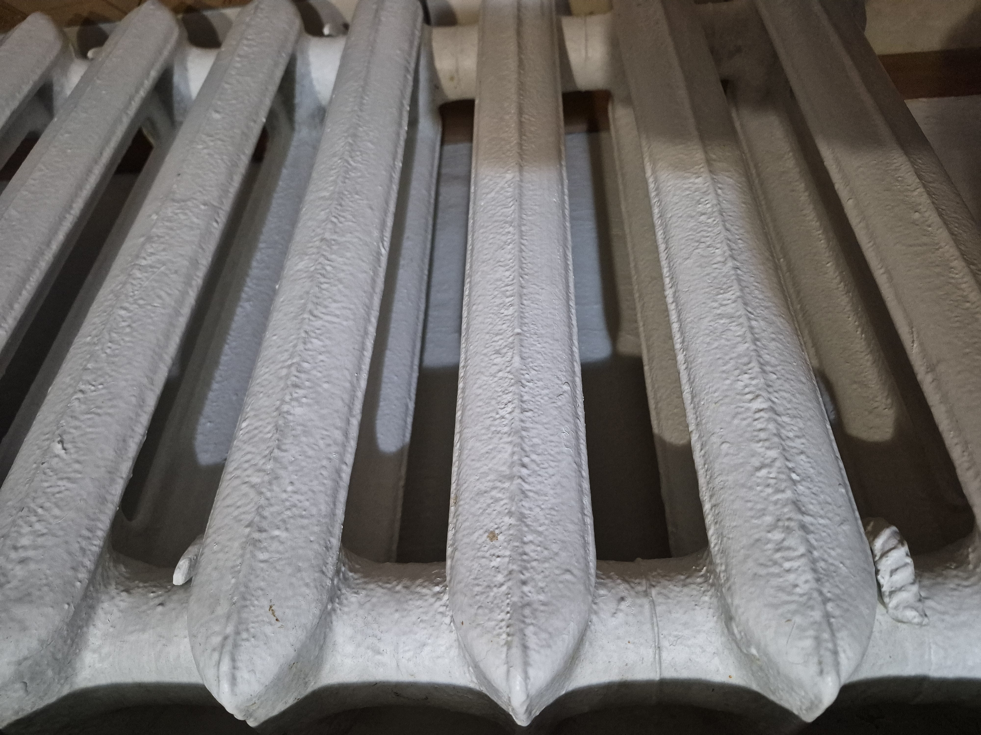 This image shows a close-up, angled view of a section of a white cast-iron radiator. The radiator has a ribbed, vertical design, with each section tapering to a point at the top and bottom. The surface of the radiator appears textured and painted, with some subtle imperfections visible. Shadows are cast between the radiator sections, suggesting a light source from above and to the side. The background is blurred but appears to be a dark, possibly wooden surface. The overall impression is of an indoor setting, likely a room heated by the radiator. There are no people or animals visible in the image, and no discernible text. The scene is static and focuses solely on the details of the radiator. Given the location context of Uglegorsk, Russia, this radiator is likely part of a heating system in a residential or public building, and the image might have been taken during a colder season when heating would be in use. There are no indicators of the time of day or weather conditions beyond what is implied by the presence of a radiator.