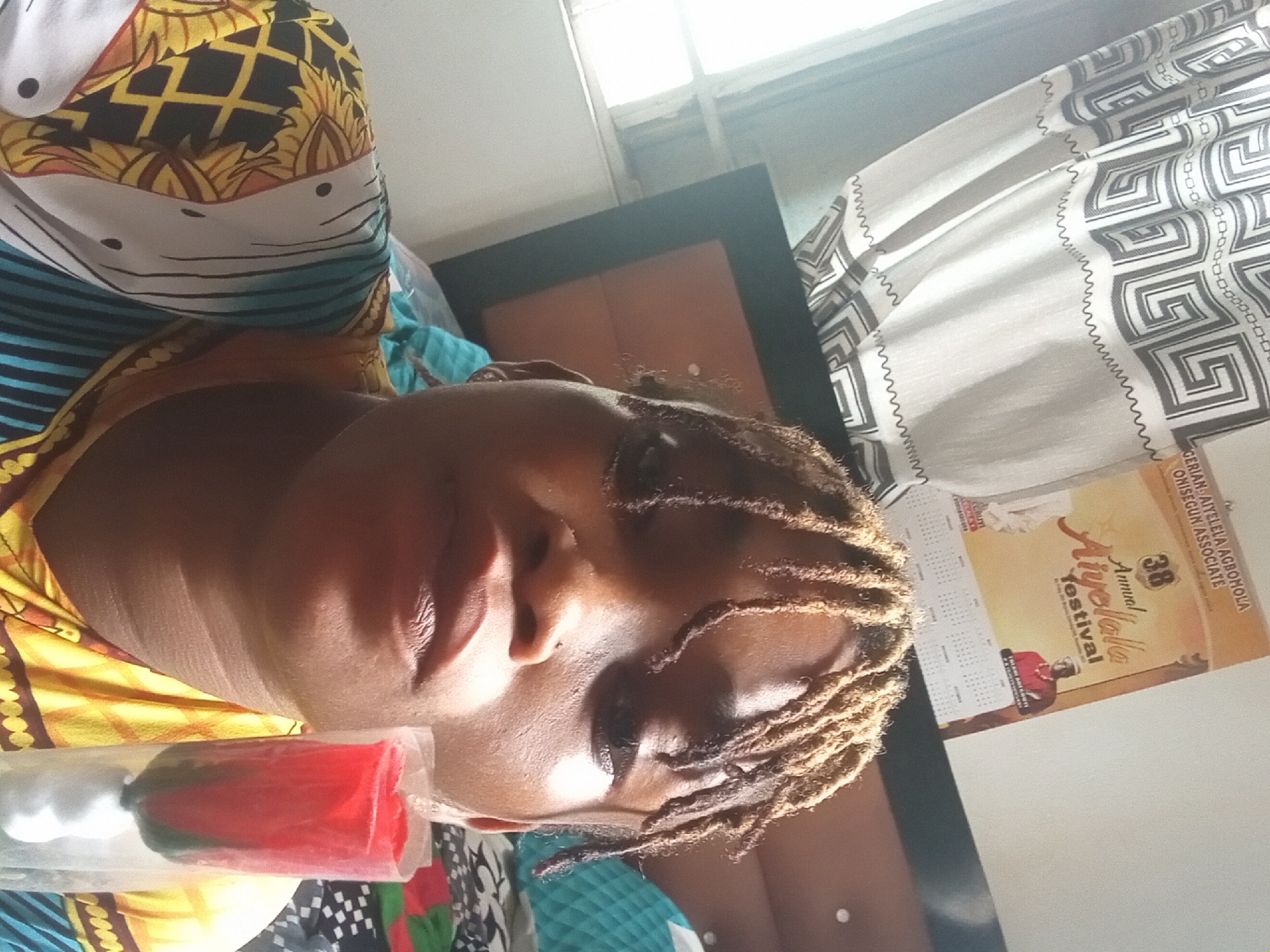A woman with blonde dreadlocks lies on a bed, her head resting on a pillow adorned with a colorful patterned cloth. She holds what appears to be a red and white object wrapped in clear plastic near her face.
The scene is indoors, evidenced by the presence of a window with white curtains featuring a geometric pattern. A calendar hangs on the wall, bearing the inscription "38 Annual Aiyekola festival." The calendar also states GERIAN: AIYELELA AGROBOLA ONISEGUN ASSOCIATE. Given that Aiyekola Festival is an annual event held in Shagamu, Nigeria, it's reasonable to suggest that this scene is likely situated in that city. A bed frame with a dark border is visible in the background.