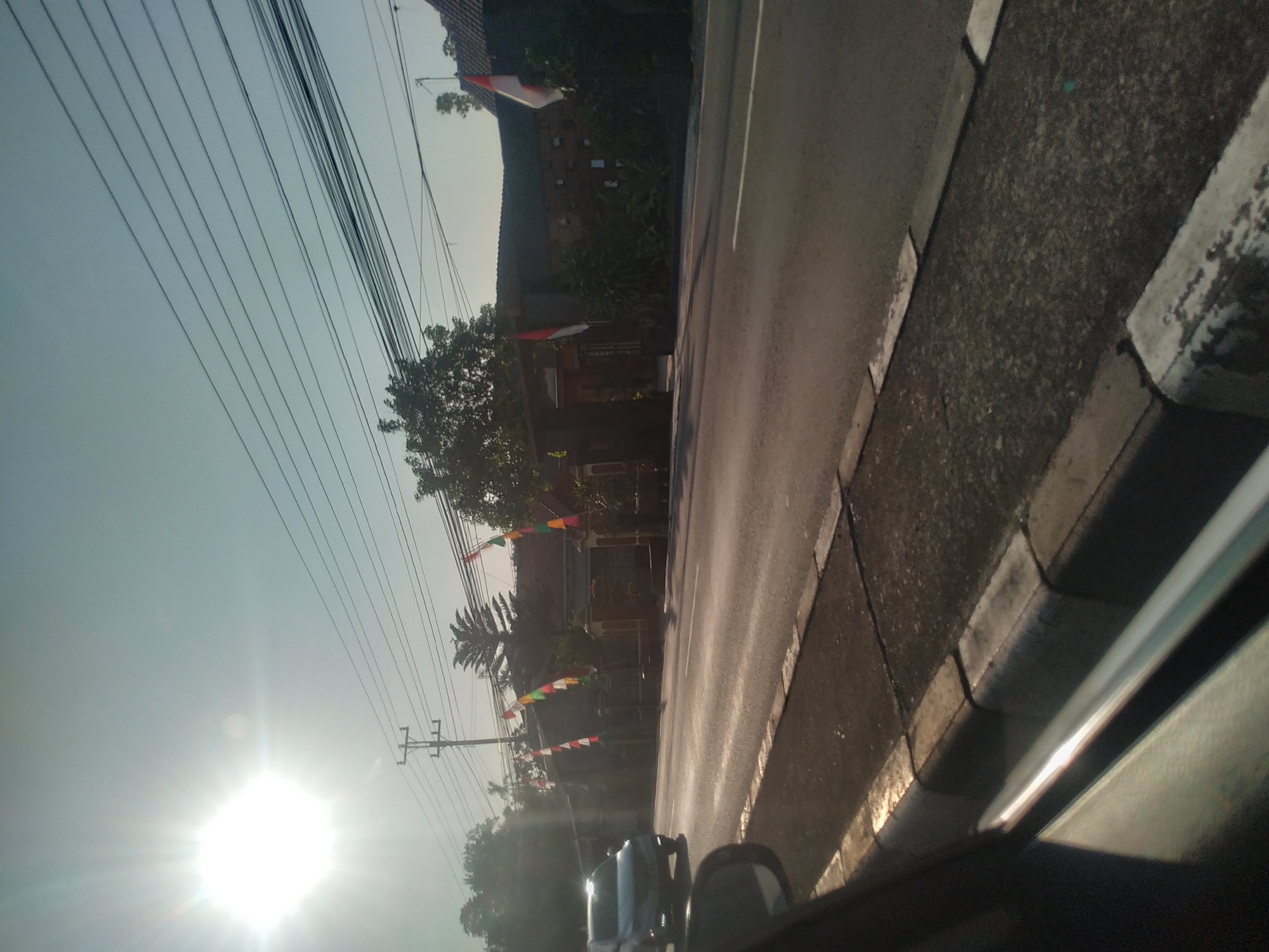 The image shows a sunlit street scene in Teras, Indonesia, likely in the morning or late afternoon given the low angle of the sun. The foreground features the edge of a road with a concrete curb and asphalt pavement. A portion of a car's side mirror and window frame is visible on the left, suggesting the photo was taken from inside a vehicle.

The road stretches into the distance, with a white line marking the center. On the right side of the road, there are several houses with traditional Indonesian architecture, featuring tiled roofs and brick or stucco walls. Red and white flags, likely Indonesian flags, are displayed at some of the properties, along with colorful triangular pennants strung along poles. Lush green trees and vegetation line the roadside and surround the houses.

Overhead, a network of power lines crisscrosses the sky, supported by utility poles. The sun is a prominent feature, casting a bright glare and creating lens flare effects in the upper left quadrant of the image. A vehicle, possibly a silver SUV, is driving on the road in the middle distance, partially obscured by the sun's glare. The overall impression is a tranquil, everyday scene in a residential area of Teras, Indonesia.