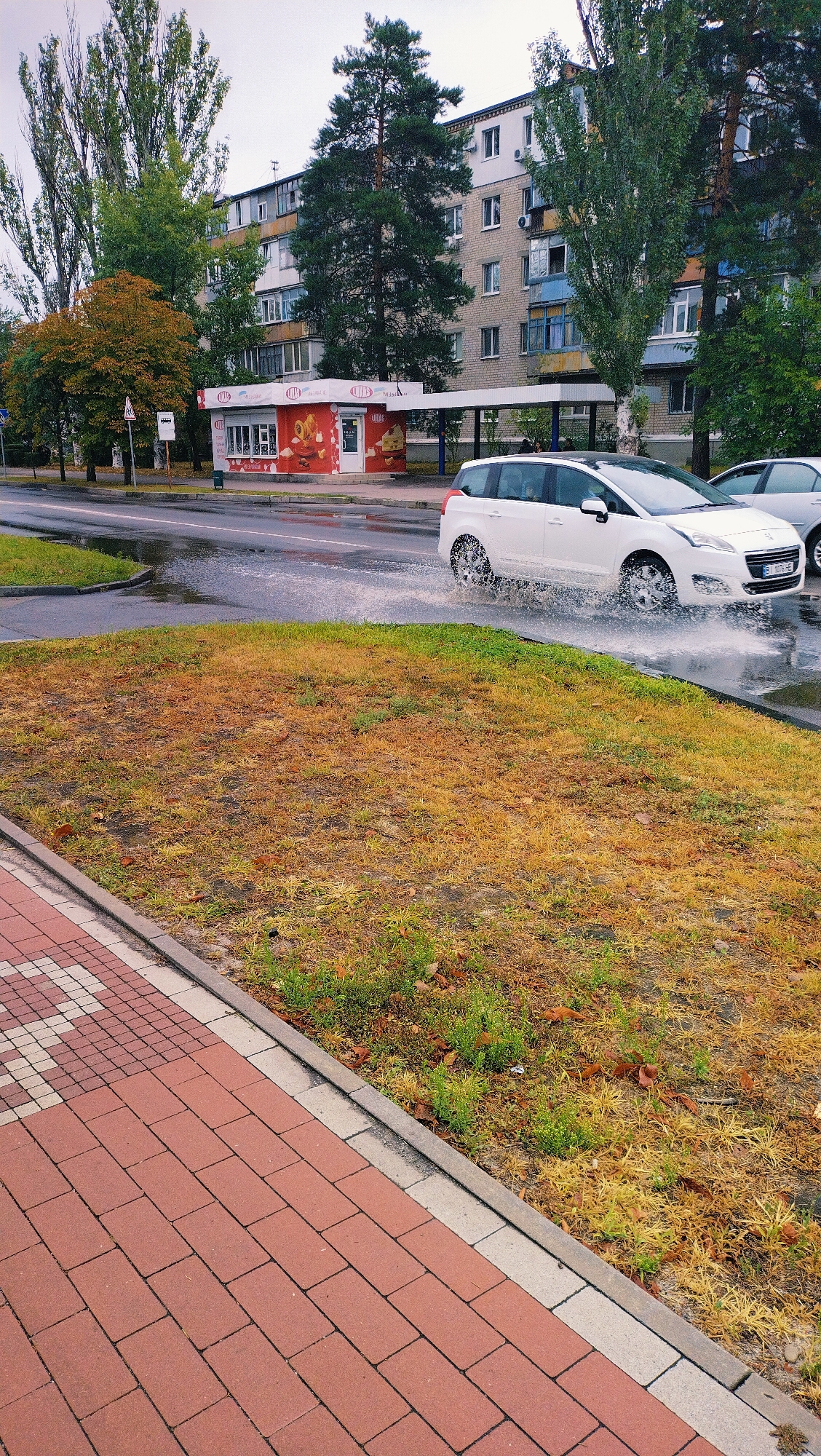 A white car is driving through a puddle on a street in Horishni Plavni, Ukraine. The car is kicking up a lot of water. The street is wet and there are puddles on the ground. There is a sidewalk with red bricks next to the street. In the background, there are apartment buildings and trees. The sky is cloudy.