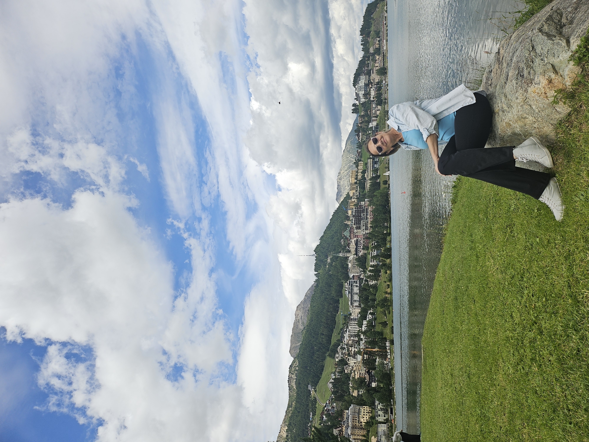 A woman is sitting on a rock in front of a lake, with a town and mountains in the background. The woman is wearing a light blue shirt, a white unbuttoned shirt, black pants, and grey sneakers. She has sunglasses on her head. The lake is calm and reflects the sky. The town is built on a hillside, with buildings of various colors and sizes. The mountains behind the town are covered in trees. The sky is blue with white clouds. The overall scene is picturesque and serene.
