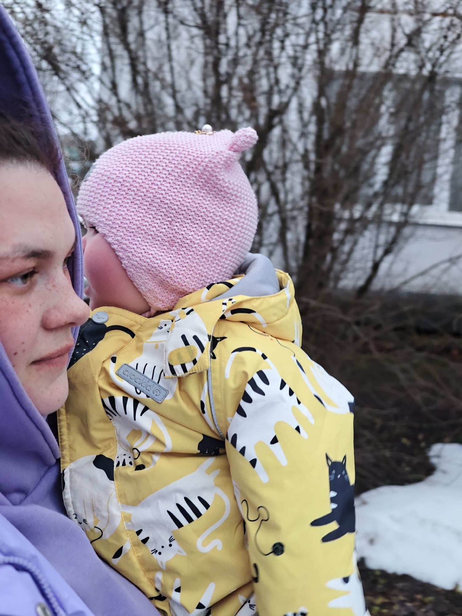 The photo shows a woman holding a baby wearing a pink knit hat and a yellow jacket with white and black cat patterns. The woman is wearing a light purple hoodie. They are standing outside, possibly in the city of Уральское, Russia, indicated by the location tag. The background includes bare trees, some greenery, and a patch of snow. A building with a window is also visible in the background.