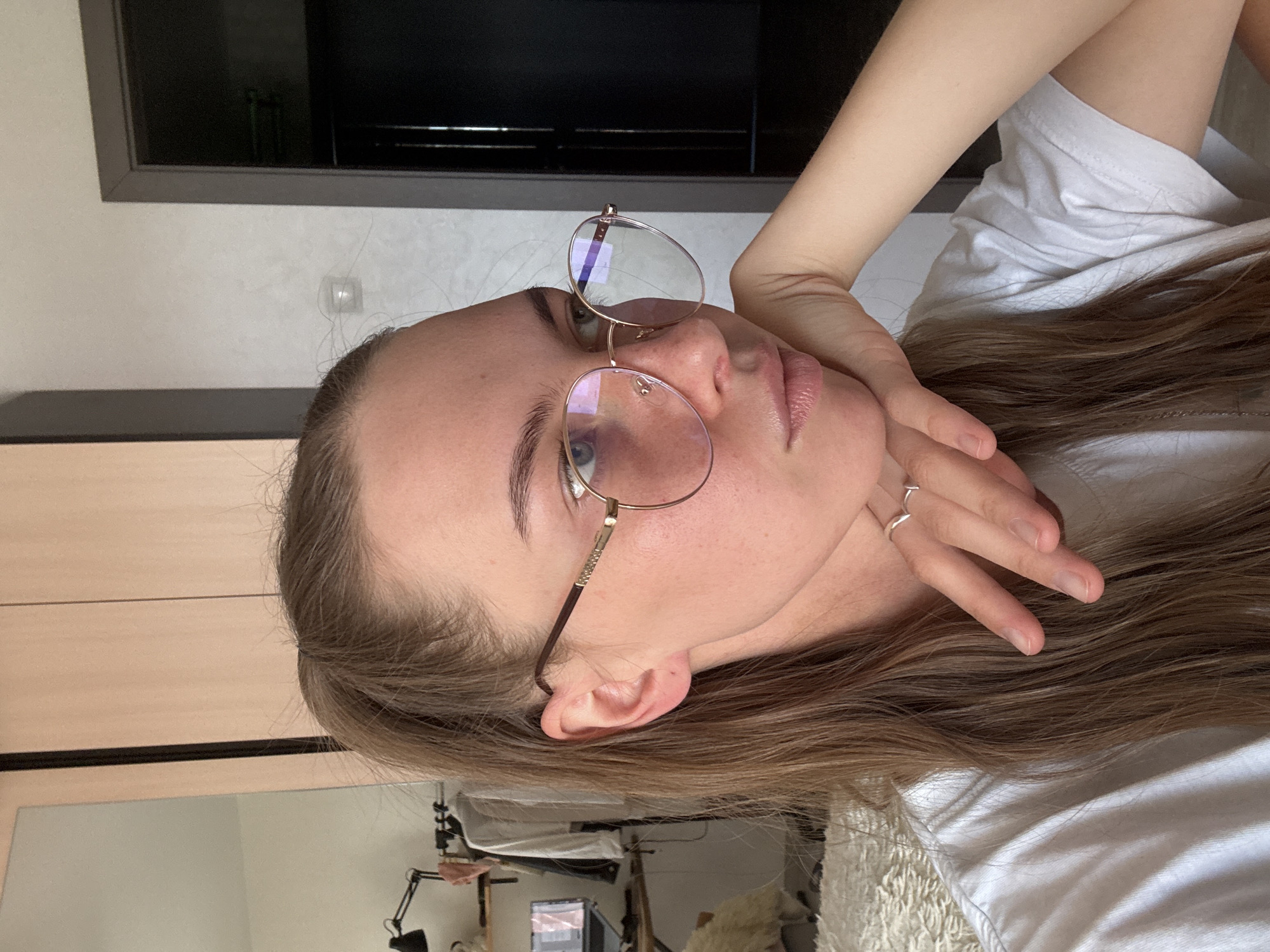A young woman with long, brown hair pulled into a ponytail wears round, gold-rimmed glasses. She is looking to her right, with her chin resting on her left hand. Her right hand is also visible, with fingers gently resting on her chin and cheek. She is wearing a white t-shirt. The background shows a closet with light wood doors and a mirror reflecting part of a room with a desk and a lamp. The scene appears to be an indoor setting, possibly a bedroom or studio apartment. There are no explicit indicators placing the image in the city of Perm, Russia.