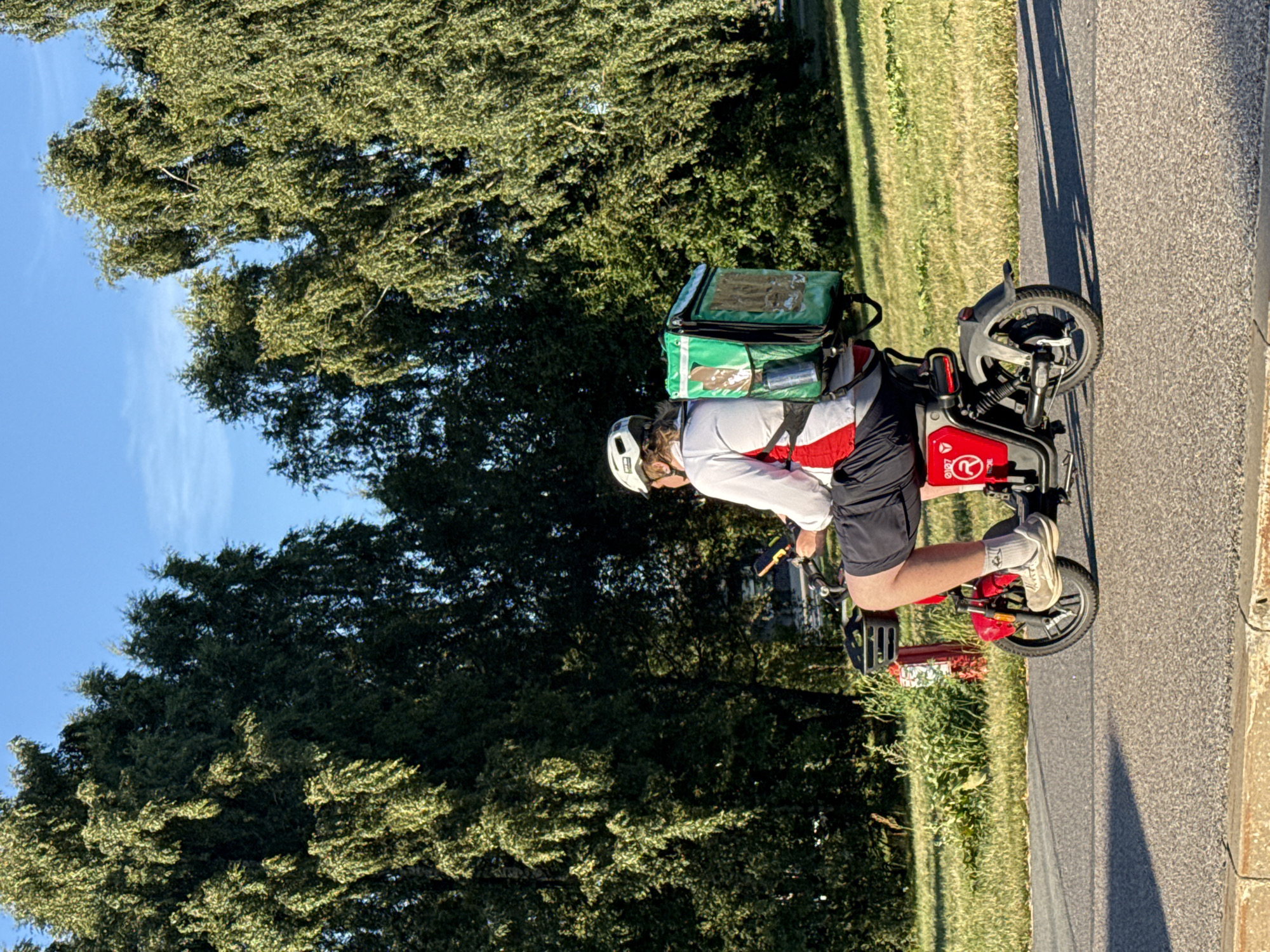 The image depicts a person riding a red electric scooter on a paved road. The individual is wearing a white helmet, a white and red jacket, black shorts, and grey socks with sneakers. They are also carrying a large green insulated delivery bag on their back, suggesting they are a food delivery worker. The scooter has a black basket in front and a red body with white logos. The scene appears to be set in an urban environment, possibly in Tallinn, Estonia, given the presence of lush green trees and a clear blue sky, indicative of a pleasant day. The road is bordered by a grassy area with more trees and sunlight casting shadows on the pavement.