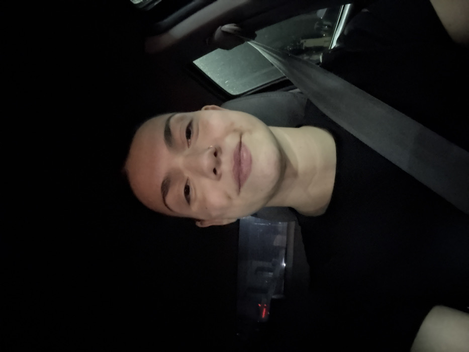 A person with fair skin and short dark hair smiles at the camera. They wear a black shirt and a seatbelt. The image appears to be taken inside a car at night. A small nose ring is visible. Part of the car's interior, including the ceiling and window, can be seen in the background. The lighting is dim, creating a high-contrast effect.