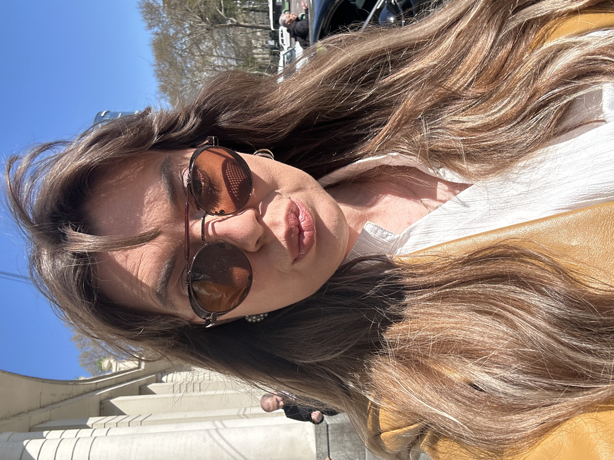A woman with long brown hair is wearing brown sunglasses, a white shirt, and a yellow jacket. She is outdoors on a sunny day, possibly on or near a set of stairs. In the background, there are trees, a building, and other people. The location is likely in Tbilisi, Georgia.