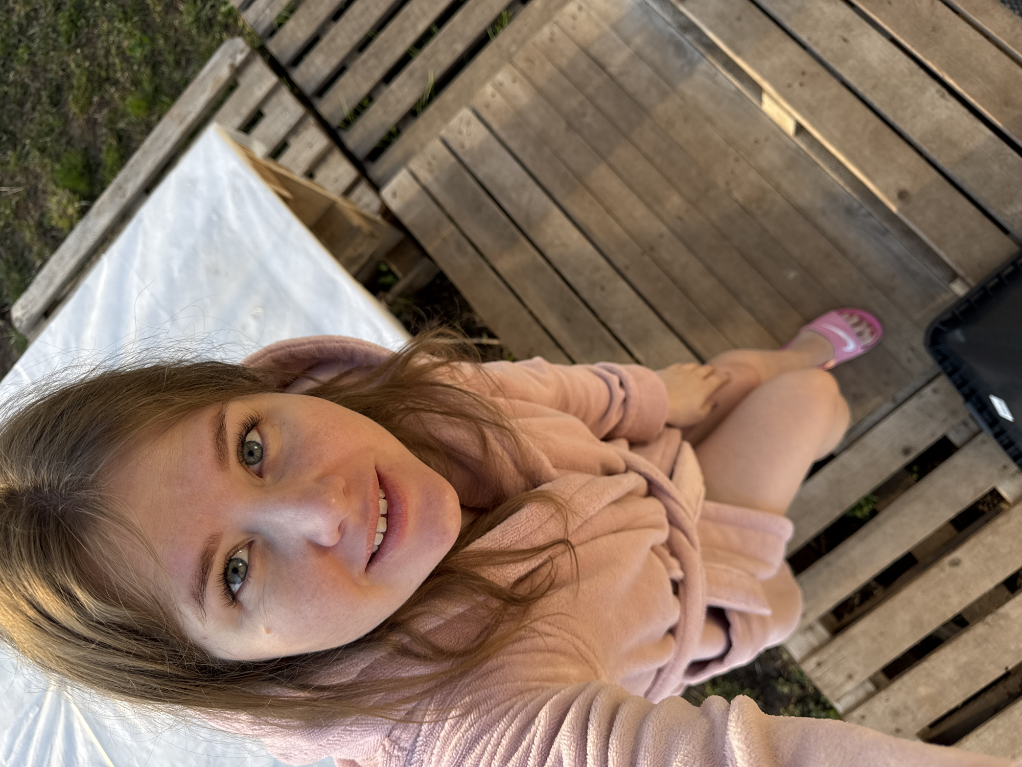 A young woman is captured in a relaxed outdoor setting, viewed from an overhead, tilted perspective. She has long, light brown hair framing her face and striking blue-green eyes, looking directly at the camera with a gentle, contented smile. She is wearing a soft, light pink robe, possibly made of fleece, which drapes around her body. Her left foot is visible in the lower right, clad in a vibrant pink slide sandal featuring a distinct white Nike swoosh logo.
The background consists of a rustic outdoor environment. A significant portion is covered by large, light-colored wooden pallets or decking, creating a textured surface. Between the slats of this wooden structure, glimpses of green grass and foliage can be seen. To the left, a large white, translucent sheet or covering, possibly part of a greenhouse or protective structure, is present. The natural lighting indicates it is daytime, suggesting a pleasant and calm atmosphere. The woman appears to be posing for a selfie or casual photograph, exuding a sense of leisure and contentment in what appears to be a farm or rural setting in Ферма, Russia.