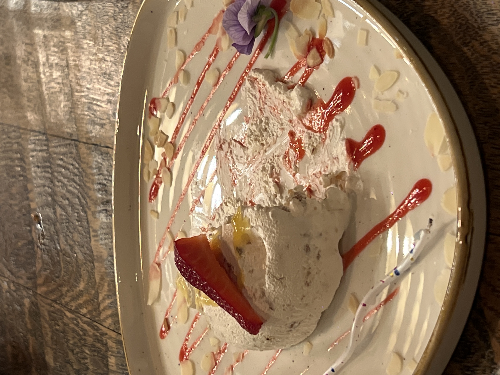 A close-up, top-down shot captures a plated dessert resting on a wooden table. The dessert consists of a light-colored, textured base topped with a dollop of what appears to be whipped cream, adorned with a slice of strawberry, a purple flower, and scattered almond slivers. A thin, reddish-brown sauce is drizzled across the plate in an artistic fashion, and there are two candles. The presentation suggests the dessert may be served in a restaurant or cafe, potentially in Yerevan, Armenia.