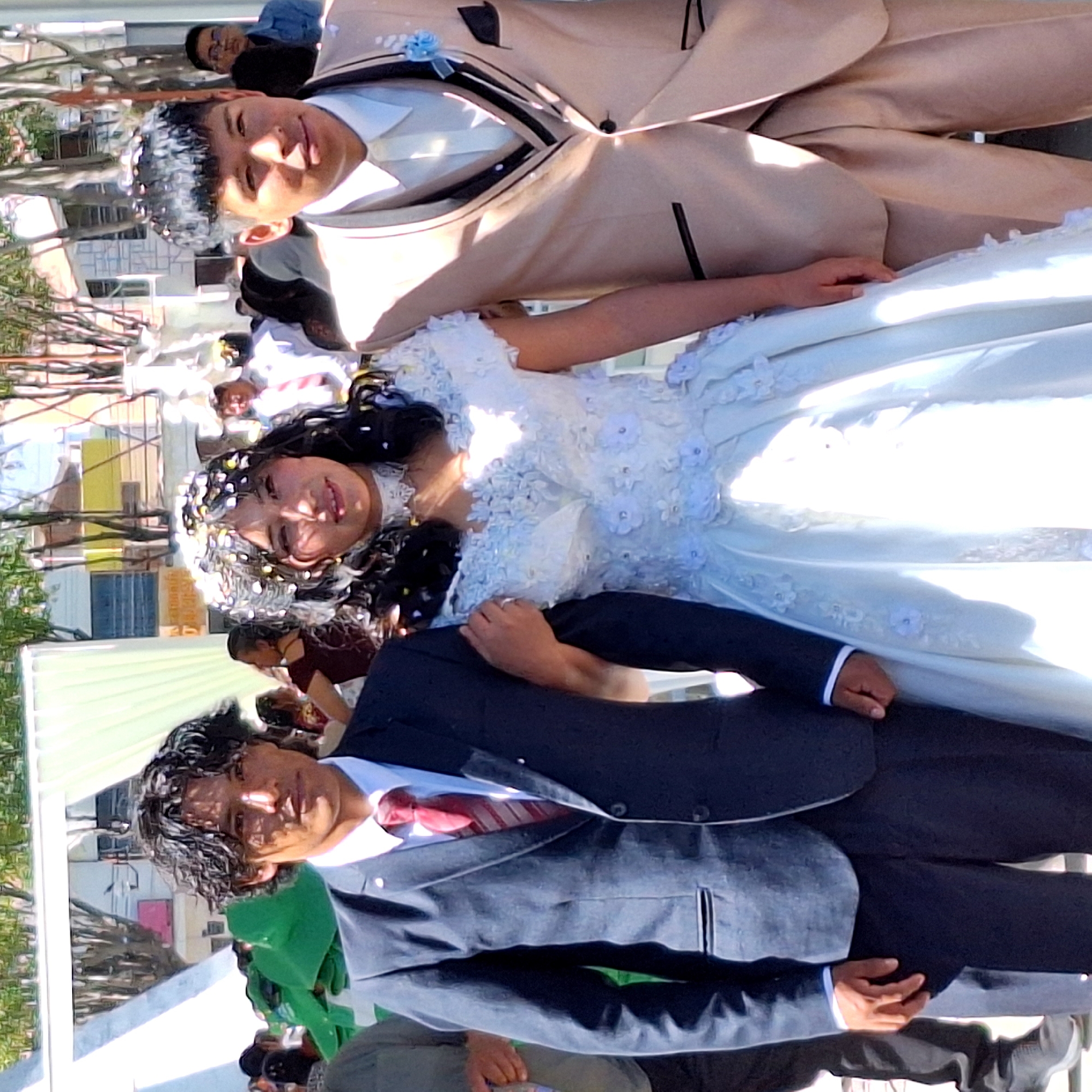 A medium shot captures a bride and groom, potentially after a wedding ceremony, in what appears to be Cerro Colorado, Peru. The bride wears a white wedding dress with floral accents. Her hair is adorned with decorative pieces. The groom wears a dark suit with a red tie. They stand outdoors, and additional people and structures are visible in the background, suggesting an urban environment.