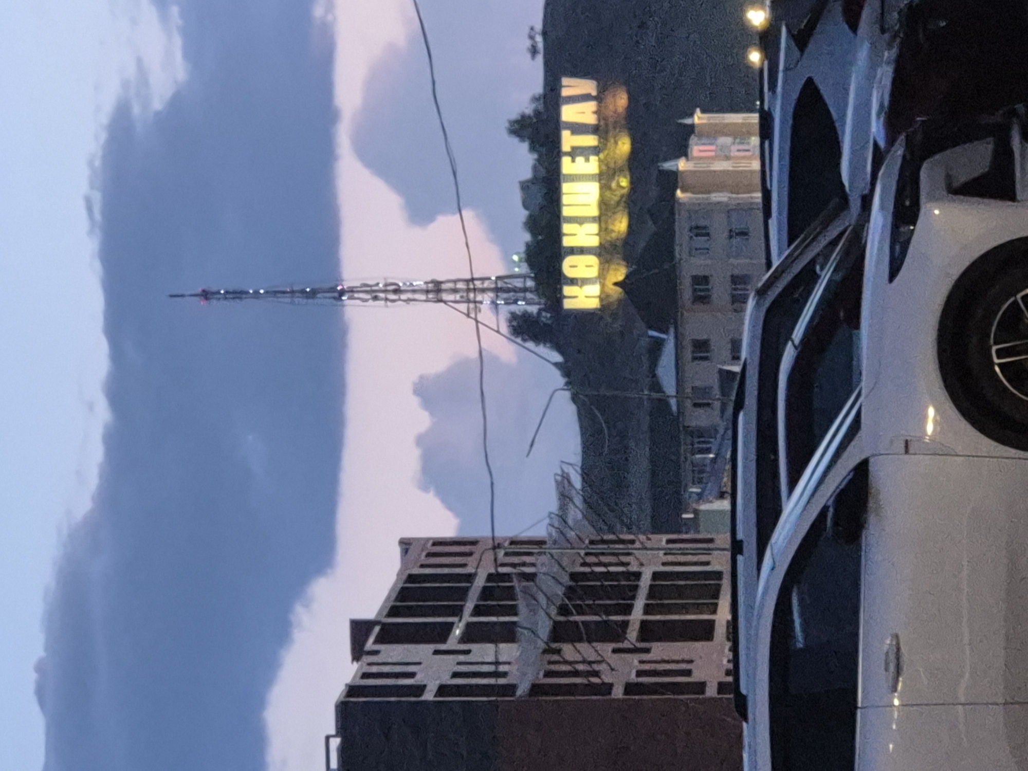 The image depicts a twilight scene in Kokshetau, Kazakhstan. In the foreground, several modern white SUVs are parked in a lot. Behind the vehicles, an unfinished multi-story building with exposed concrete structures is visible. Further in the background, on a hill, a large illuminated sign spells out "КОКШЕТАУ" in yellow lettering. A tall communication tower stands behind the sign, and several buildings with lit windows are scattered across the hillside. The sky above is a blend of deep blue and purple, with distinct cloud formations. The overall atmosphere suggests an urban environment at dusk.