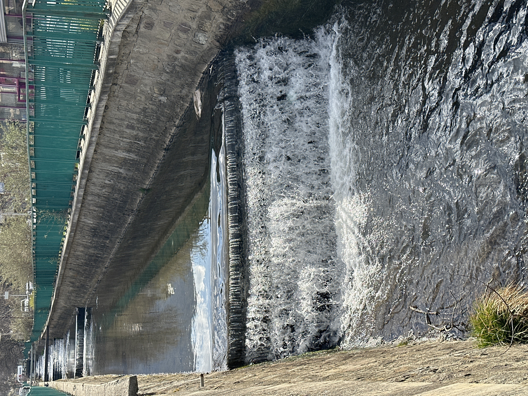 The image displays a section of the Perlovska River in Sofia, Bulgaria, characterized by its canalized concrete and stone construction. In the foreground, water cascades over a concrete weir, forming a turbulent white waterfall. Further upstream, at least two more smaller weirs are visible, creating a stepped series of cascades. The water in the canal reflects patches of blue sky and bare tree branches. The right bank features a retaining wall composed of concrete and rough-hewn stone blocks, topped by a continuous dark teal metal railing with vertical slats. Beyond this railing, sparse, bare deciduous trees and various urban structures are discernible, including buildings with dark windows and some purple-red architectural elements, alongside distant vehicles on what appears to be a street, one bearing a faint "Speedy" logo. The left bank is a simpler concrete wall, with isolated tufts of green and brown vegetation clinging to its base near the water level. The scene is illuminated by bright daylight, causing prominent glints and reflections on the water's surface. No individuals are present in the frame.