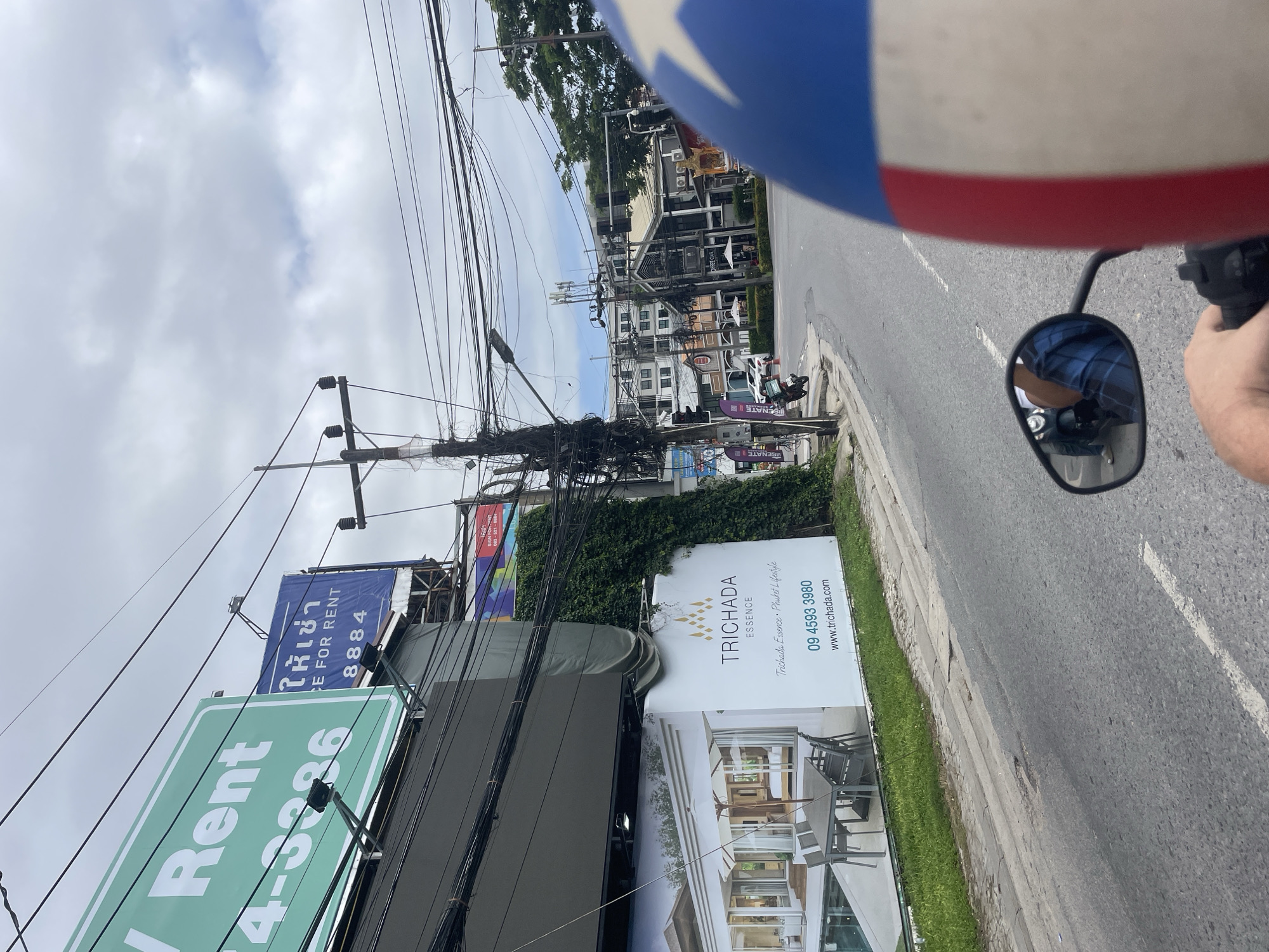 The image captures a street scene in Choeng Thale, Thailand, from the perspective of a person riding a scooter. In the immediate foreground, the top of a blue, white, and red helmet is visible on the right, along with the rider's hand gripping the scooter handlebar. The scooter's left rearview mirror reflects a partial view of the rider, dressed in a blue plaid shirt.
The road ahead is asphalt with a faded white dashed line in the center. To the left of the road, a grassy verge precedes a large white billboard for "TRICHADA ESSENCE," featuring an image of a modern house with a pool and outdoor dining area. The billboard includes text "Trichada Essence • Phuket Lifestyle," a contact number "09 4593 3980," and a website "www.trichada.com." Above this, a tall utility pole is heavily laden with a dense tangle of black electrical and communication wires. Further down the street on the left, a dark-colored moped is parked near the sidewalk.
Beyond the "TRICHADA ESSENCE" billboard, an adjacent building facade is covered in green ivy. Above these, multiple billboards are visible: a large, dark rectangular one, a partially obscured green one displaying "Rent" and numbers "4-3386," and a blue billboard with Thai script "ให้เช่า" (for rent) and English text "CE FOR RENT" and "8884."
The road curves slightly to the right in the midground, revealing an intersection with traffic lights. Several multi-story buildings line the street, featuring various architectural styles. One prominent building on the right appears to be "COFFEE CLUB" with outdoor seating and umbrellas. Another utility pole with numerous wires is visible on the right side of the street. The sky is overcast with a mix of grey and lighter clouds. Green trees are visible in the distance, behind the buildings on the right.