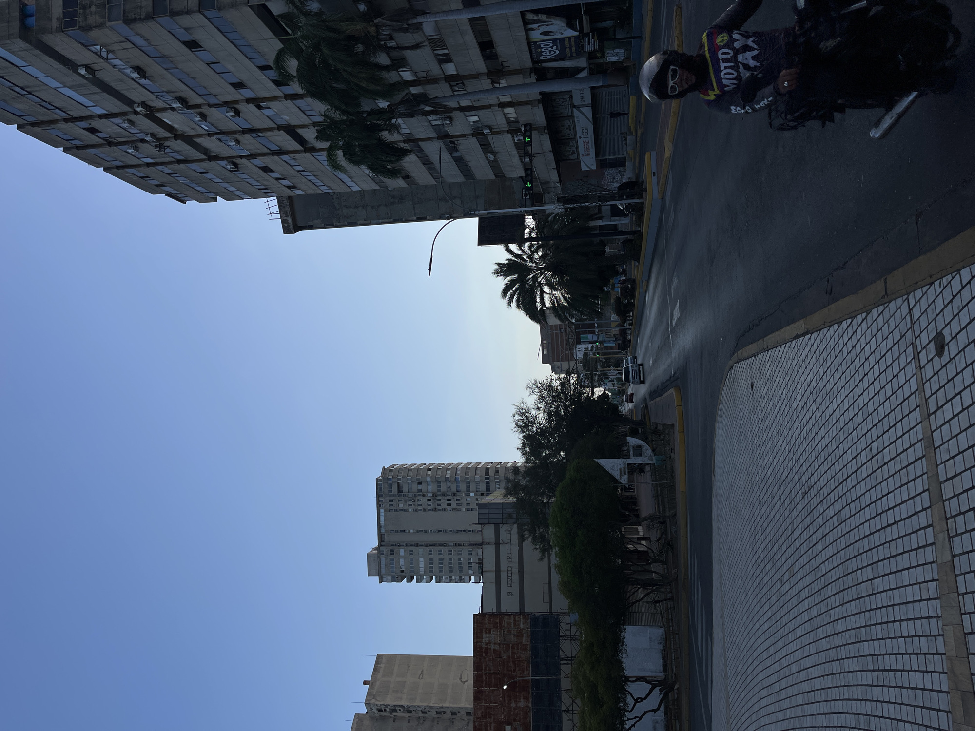 The image depicts a street scene in Porlamar, Venezuela. A tall, older building with numerous windows is on the left side of the street. A motorcycle rider with a helmet and a "Moto Taxi" shirt is visible on the right side of the street. The street surface is paved, and there is a tiled sidewalk. Other buildings and palm trees are also in the scene.