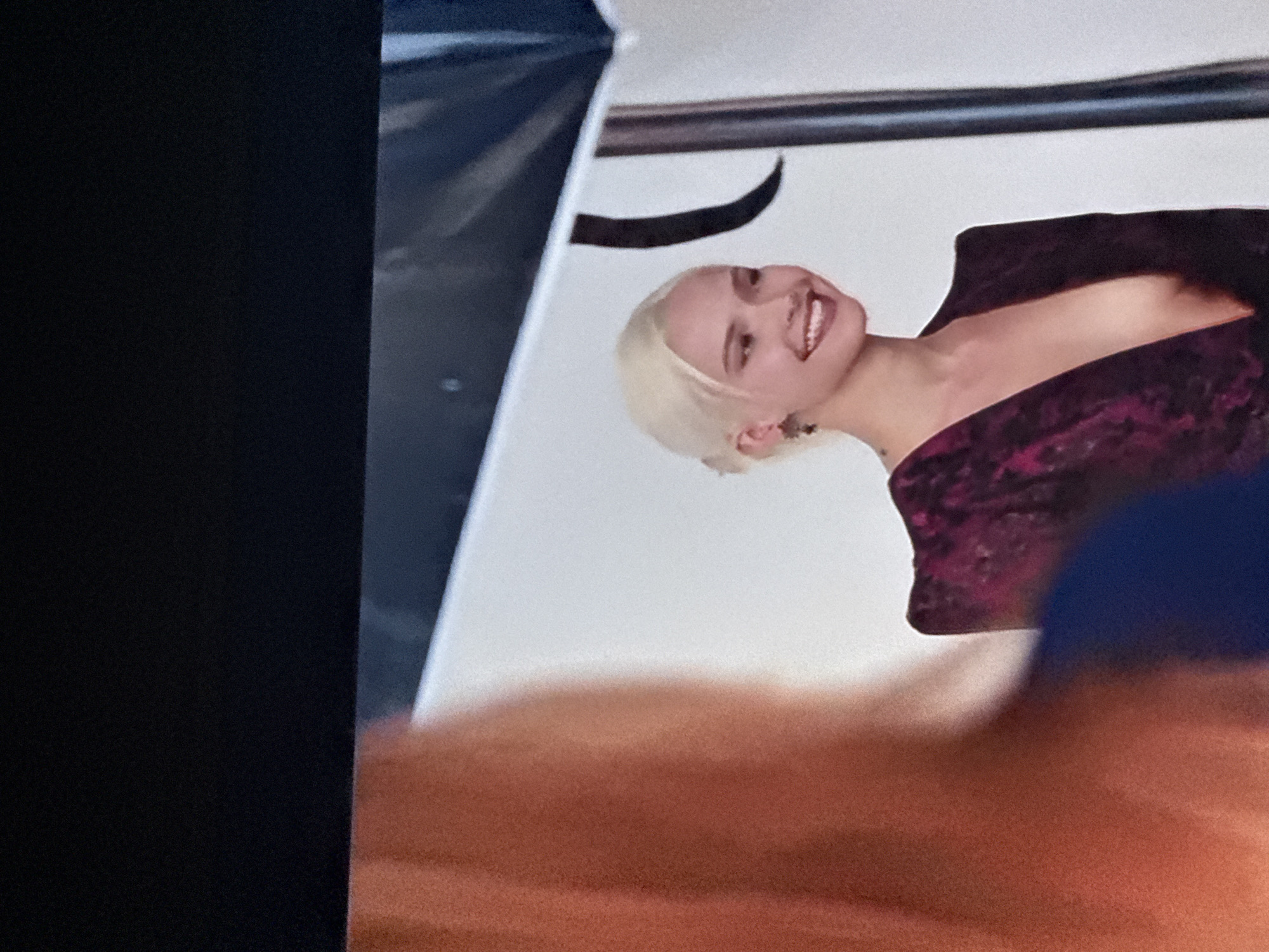 The image shows a person with blonde hair and a purple floral-patterned garment. The person is smiling and appears to be posing in front of a white backdrop. A portion of the image is obscured by an out-of-focus object in the foreground. The upper left corner is black, possibly part of the screen from which the image was taken. There's no indication of Mytishchi, Russia, in the scene.