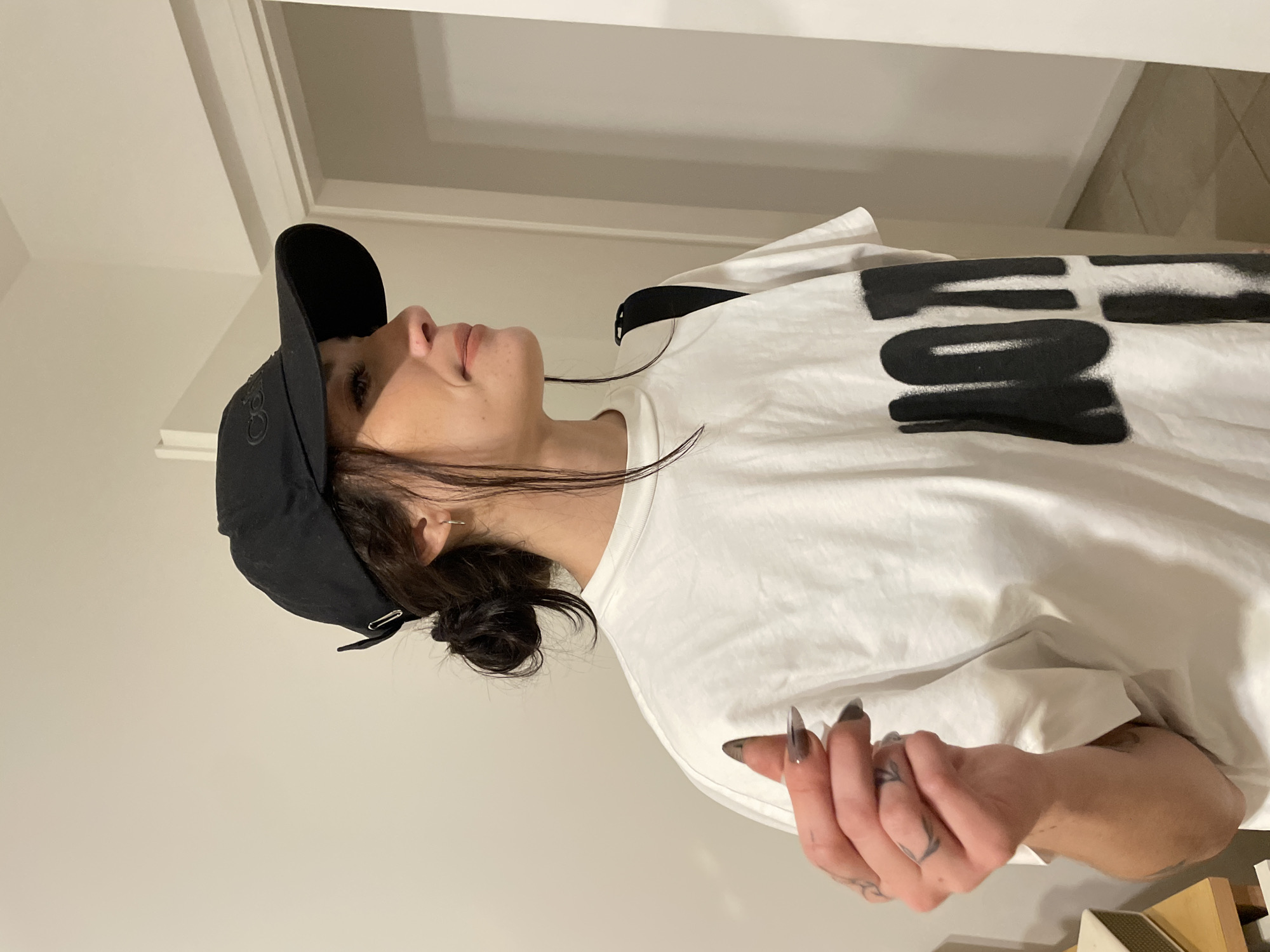 A woman wearing a black baseball cap and a white t-shirt with large black lettering stands indoors. The walls are light-colored, and there is a doorway in the background. The location appears to be inside a home or apartment. There is no indication of it being in the city of Buenos Aires, Argentina.