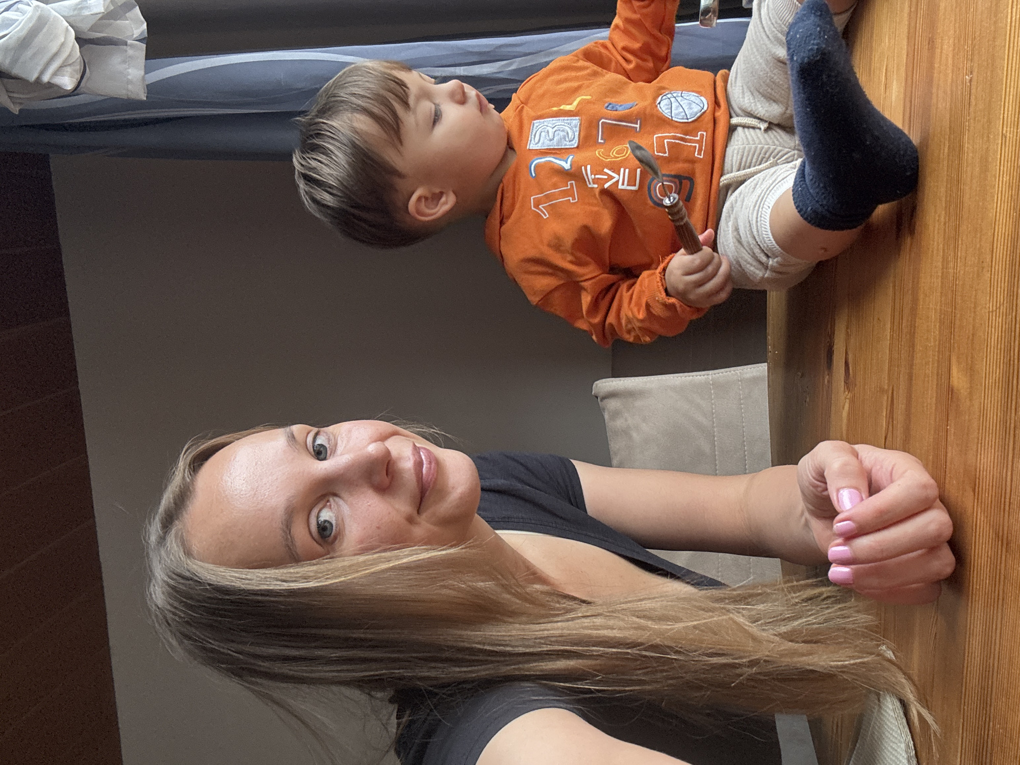 In Überherrn, Germany, a woman with blonde hair and a black t-shirt leans over a wooden table. She has light blue eyes and is smiling. Her right hand, with pink nail polish, rests on the table. To her right, a young child wearing an orange hoodie with numbers and a basketball graphic, and light-colored pants, sits on the table. The child holds a small metal utensil in their right hand and wears a dark blue sock on their left foot. A blue curtain is visible in the background. The scene is captured in natural light.