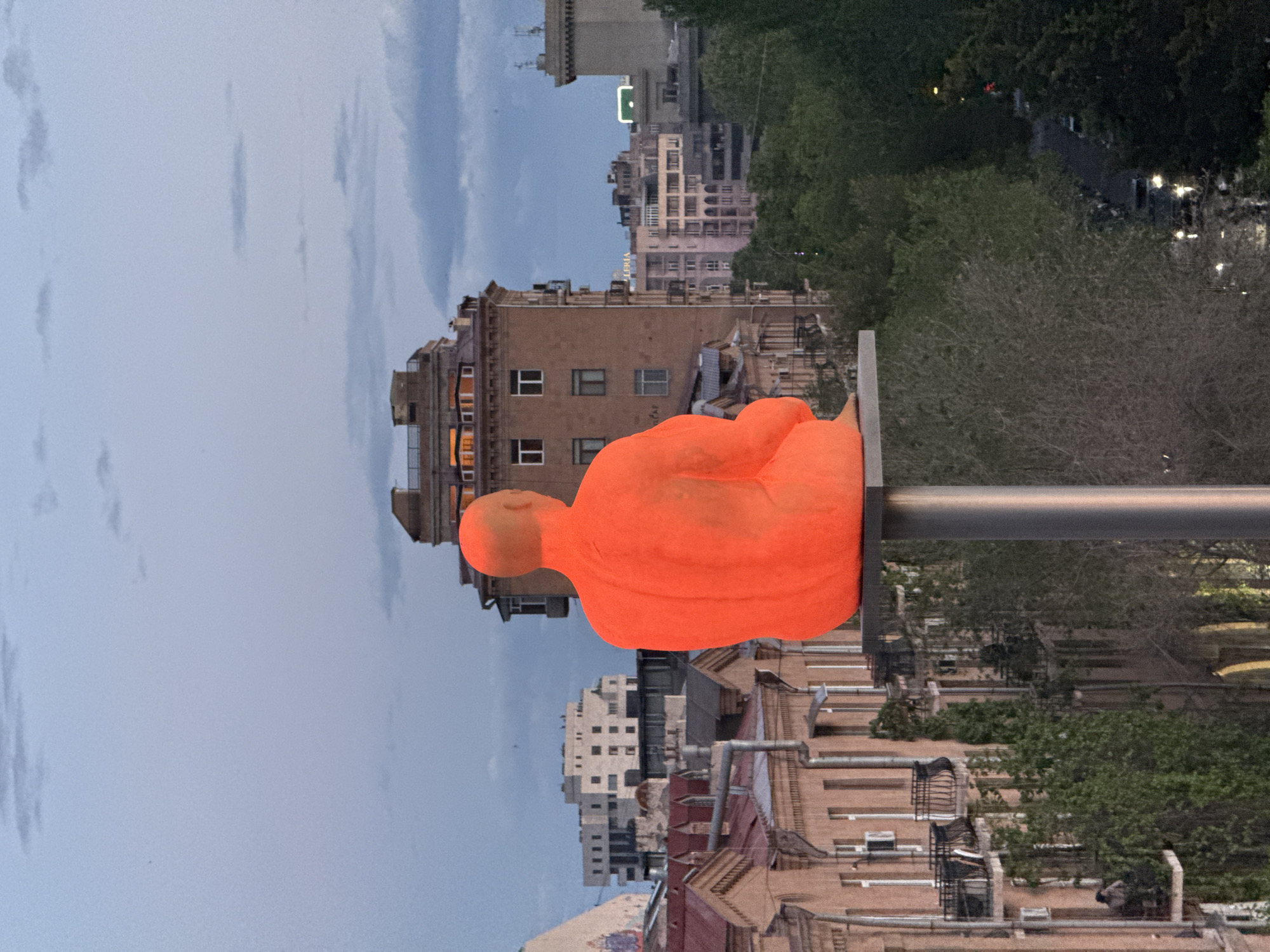 The image shows an orange statue of a seated figure, possibly meditating, placed atop a metal pole against a backdrop of buildings and trees. The setting appears to be urban, featuring a mix of older, multi-story structures with visible architectural details, interspersed with greener areas of trees. The sky is overcast. The statue is positioned on the side of the street. Given the statue's style and placement, as well as the architectural characteristics of the buildings, the location is most likely in Yerevan, Armenia.
