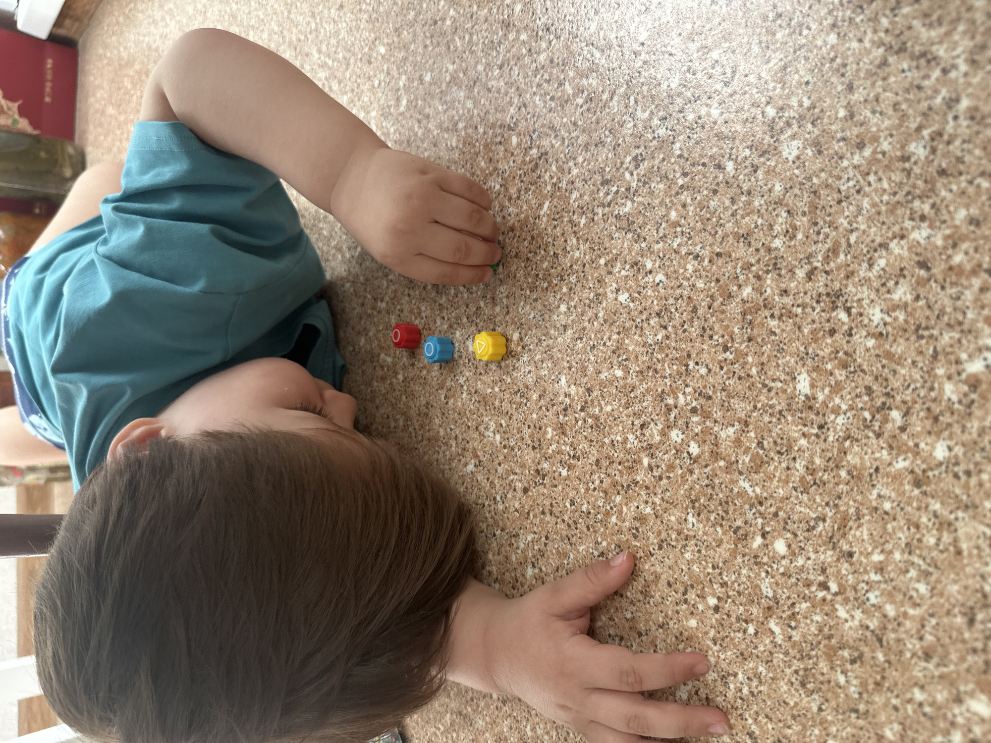 A young child is lying on a speckled brown and beige floor, looking down at three small, colorful plastic building blocks. The blocks are arranged vertically, with a red block at the top, a blue block in the middle, and a yellow block at the bottom. The child's head is resting on the floor, and their right hand is reaching out towards the blocks. The child is wearing a teal t-shirt. The scene appears to be indoors, possibly in a home or a play area. There are no visible indications of the specific location in Ussurijsk, Russia.