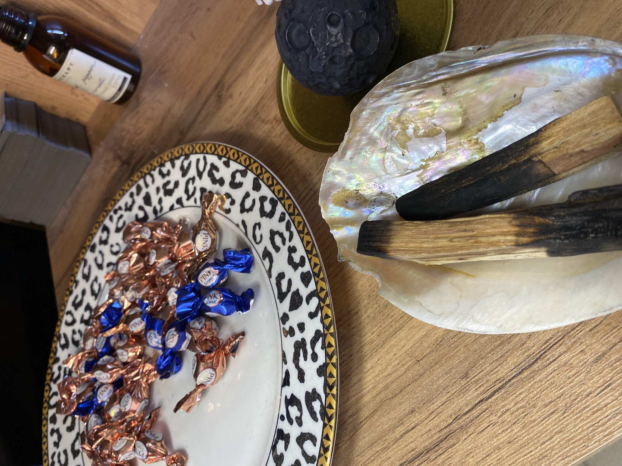 A high-angle shot shows a wooden surface with several items arranged on it. In the upper left is a stack of gray, rectangular objects and a brown glass bottle with a white label. To the right of these, a plate with a black and white animal print rim holds a pile of wrapped candies in copper and blue wrappers with the brand "PINK" visible. Adjacent to the plate is a large, iridescent seashell containing two pieces of wood, one with a charred end. A small, gray moon-shaped object sits on a round, gold-colored base between the shell and the plate. There are no people visible, and the environment suggests an indoor setting. The city of Votkinsk, Russia is not discernible from the image.