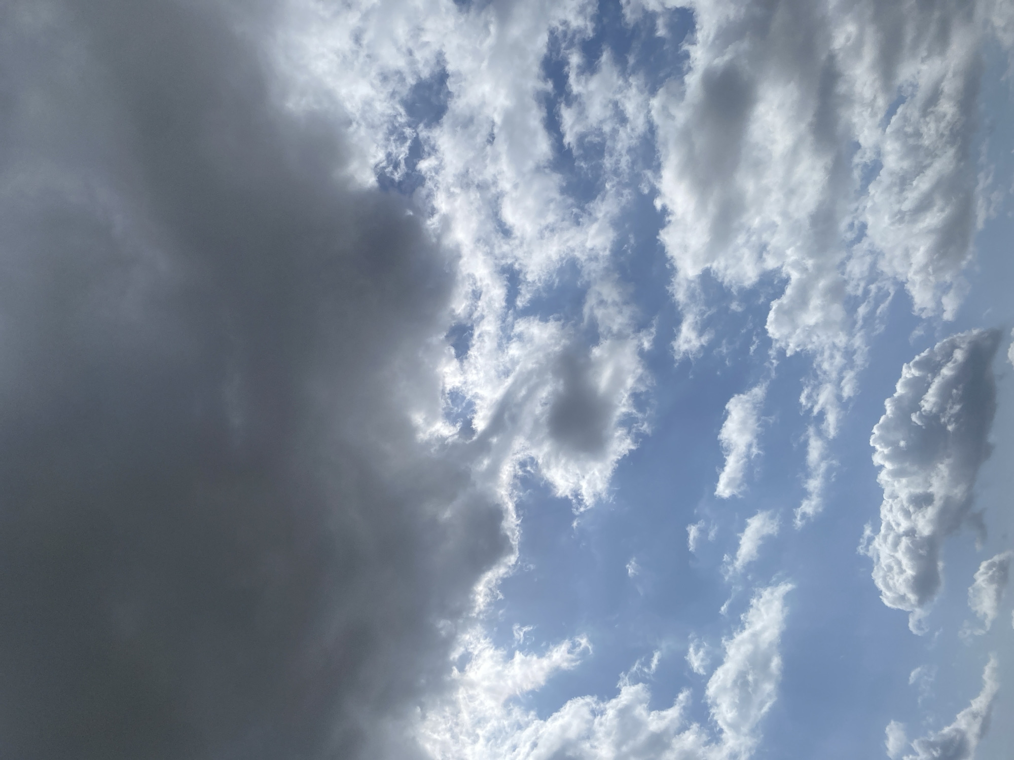 The image shows a sky filled with clouds, with a mix of dark and light formations. The upper portion of the image is dominated by large, dark, heavy clouds, suggesting an overcast or stormy condition. Below these, lighter, wispy white clouds are scattered across a blue sky. In some areas, sunlight breaks through the darker clouds, illuminating patches of the lighter clouds. The overall impression is of a dynamic sky, potentially a transition between clear weather and a storm, or vice versa. The scene is exclusively a sky view, with no visible ground elements, buildings, or people. The location in Jos, Nigeria, is not discernible from the visual information provided in the image itself.