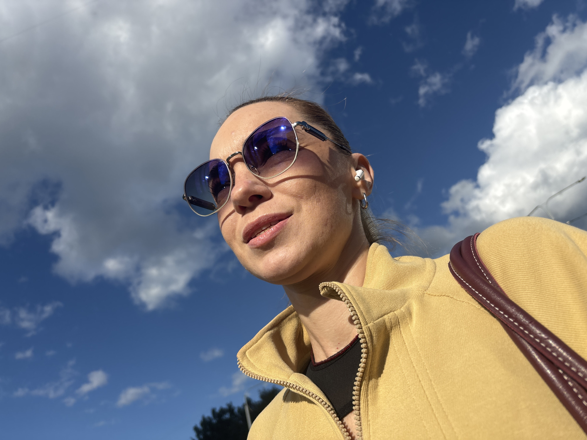 A low-angle, eye-level shot captures a woman from the chest up, looking to her left. She wears a yellow zip-up jacket over a black top, with a burgundy strap from a purse visible across her right shoulder. She has on reflective sunglasses with purple lenses and a white wireless earbud in her right ear, with a hoop earring on that ear. Her hair is pulled back, and she has a faint smile, with glossy lips. The scene is outdoors under a bright blue sky dotted with fluffy white clouds. The lighting suggests it is daytime, possibly mid-morning or afternoon. There is no visible text in the image. The background consists of the sky and clouds, with a hint of dark green foliage at the bottom left edge of the frame, suggesting an outdoor setting that could be a park or urban area.