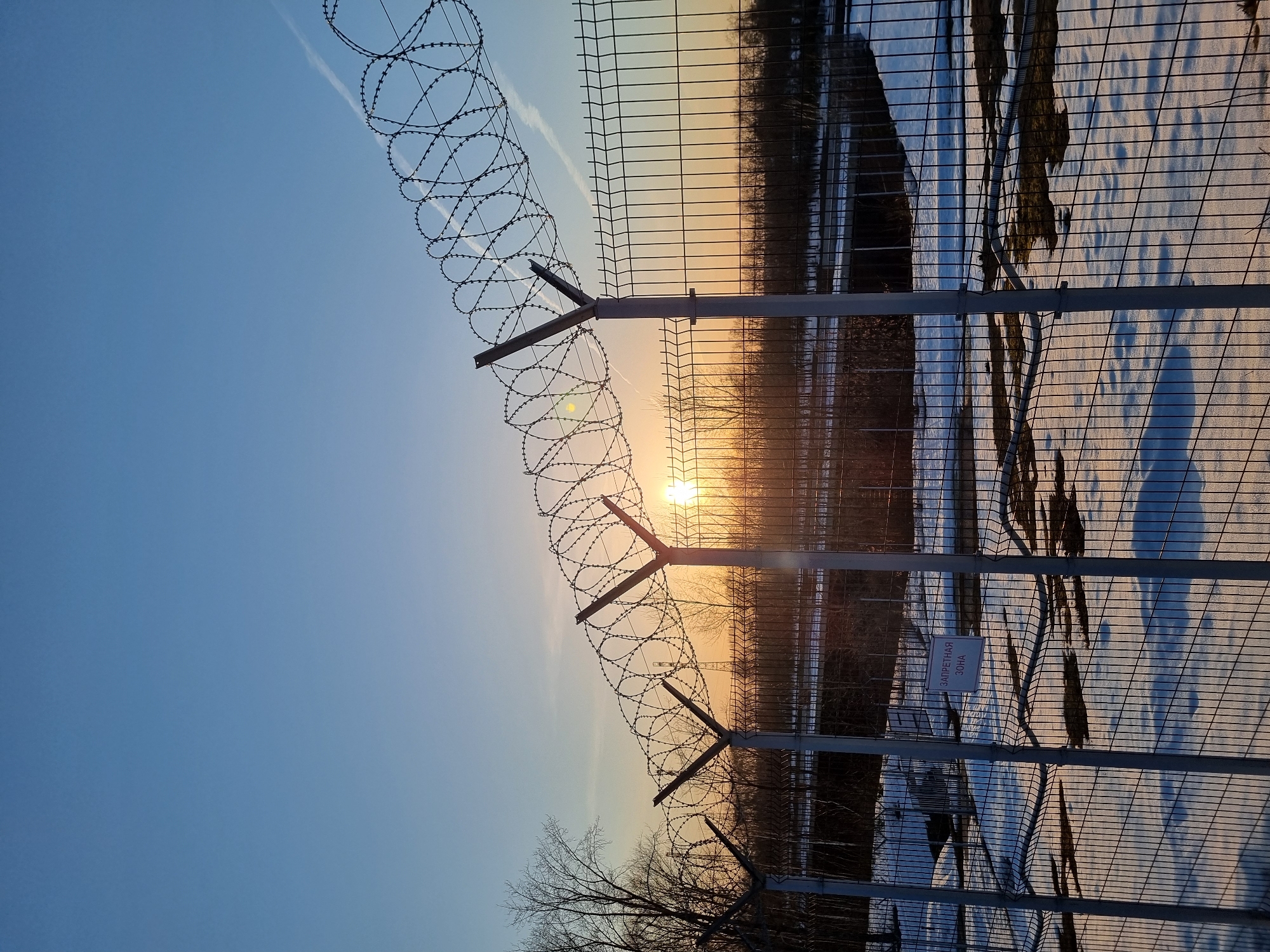 The image depicts a security fence topped with barbed wire, presumably in Chaikovsky, Russia, given the text on the sign attached to the fence. The fence is constructed of a wire mesh attached to metal posts. The barbed wire runs along the top of the fence in coiled sections. Behind the fence is a landscape with trees and snow-covered ground. The setting sun is visible in the background, creating a warm, orange glow. The sky is blue, transitioning to a lighter hue near the horizon. A contrail is visible in the upper left of the frame. The sign on the fence is in Russian and translates to "Restricted Area". The lighting suggests either sunrise or sunset. No people are visible in the frame.