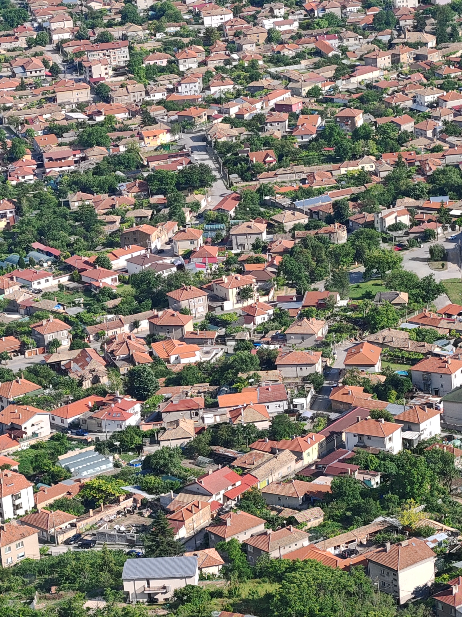 This image presents an elevated, panoramic view of a densely packed residential district within the city of Shumen, Bulgaria. The scene is characterized by numerous houses, predominantly two to three stories in height, featuring traditional red or orange terracotta-tiled roofs. Buildings exhibit a mix of architectural styles, ranging from older structures with plaster facades to more modern constructions. Extensive green foliage, including numerous trees and bushes, is interspersed throughout the settlement, often surrounding individual properties and lining the winding asphalt roads that traverse the area. Several vehicles (cars) are visible on the roads and parked near residences. Visible details include solar panels on some rooftops, a few greenhouses or poly-tunnels in yards, and an above-ground swimming pool in one garden. No individuals are clearly discernible within the frame. The perspective suggests the image was captured from a significant elevation, likely from the Shumen Plateau, looking down upon the city's urban sprawl.