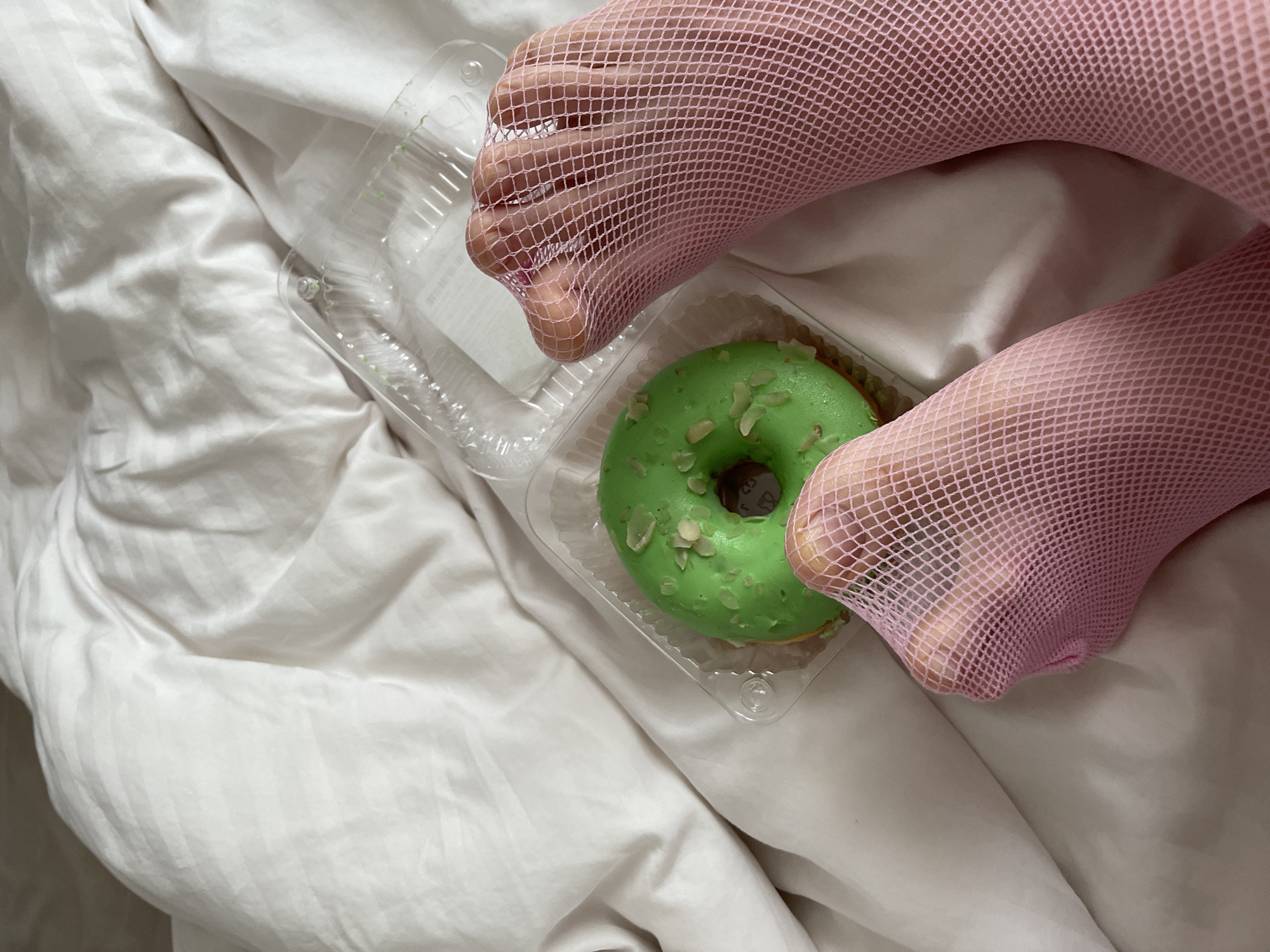 A close-up shot shows a green donut with white slivers of nuts inside a clear plastic container. The container is placed on a rumpled white sheet. Two feet, covered in pink fishnet stockings, are positioned on either side of the donut. The toes of the feet are visible through the mesh. The lighting is soft, suggesting an indoor setting with diffused light, possibly natural light from a window or soft artificial lighting. The overall impression is intimate and perhaps a bit playful, with the contrasting textures of the stocking, the sheet, and the donut creating visual interest. The focus is on the feet and the donut, implying a personal moment or a specific aesthetic. The scene is likely set in a bedroom or a similar private space, given the presence of the sheet. There is no visible text in the image.