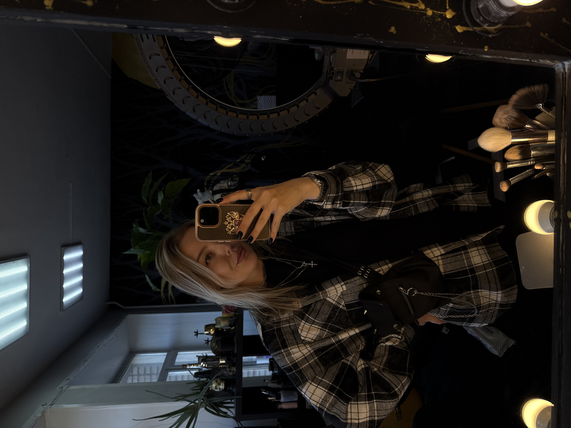 A woman takes a selfie in a makeup mirror. The mirror is surrounded by lights. Makeup brushes are visible in the upper right corner of the mirror. The woman is wearing a black top and a black-and-white plaid shirt. She is wearing a silver necklace. She has blonde hair. The phone she is using has a gold case with an emblem. In the background behind the mirror, there is a ring light and a plant. Through the mirror, it shows a room lit by fluorescent lights, with shelves, decor, and plants. Based on the shelving decor and makeup setup, this is likely a salon or studio setting in Krasnoyarsk, Russia.