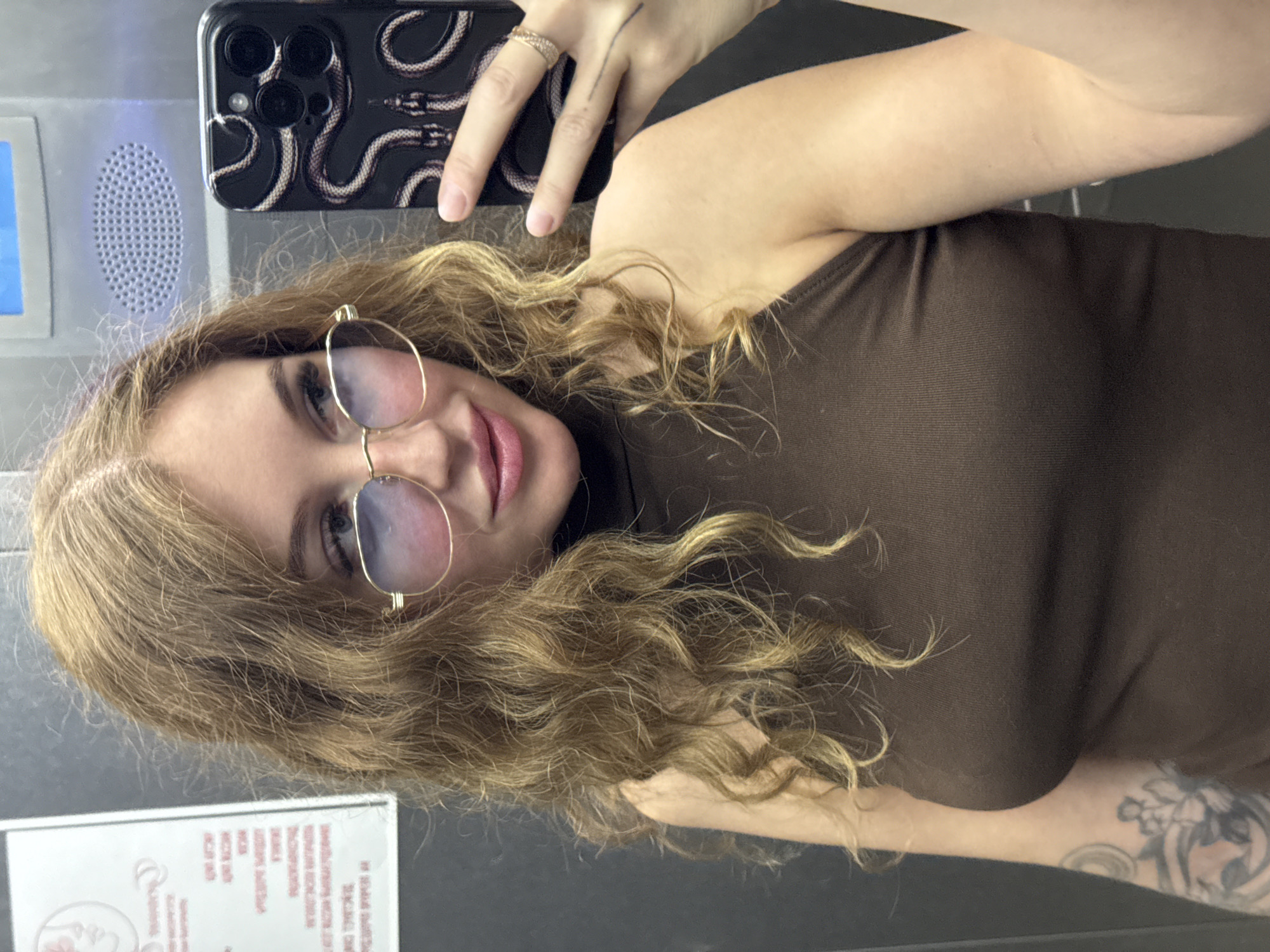 A young woman with blonde, wavy hair takes a selfie in a bathroom. She is wearing a brown, sleeveless top and gold-rimmed glasses. Her makeup includes pink lipstick and eyeliner. Her left hand, adorned with a ring, holds a smartphone with a black case featuring a snake pattern. Visible on her left arm are tattoos. The background includes a speaker grill on a light-colored panel and a dark gray wall. A portion of a document with red text is visible at the bottom of the frame, containing Cyrillic lettering. The lighting suggests an indoor setting.