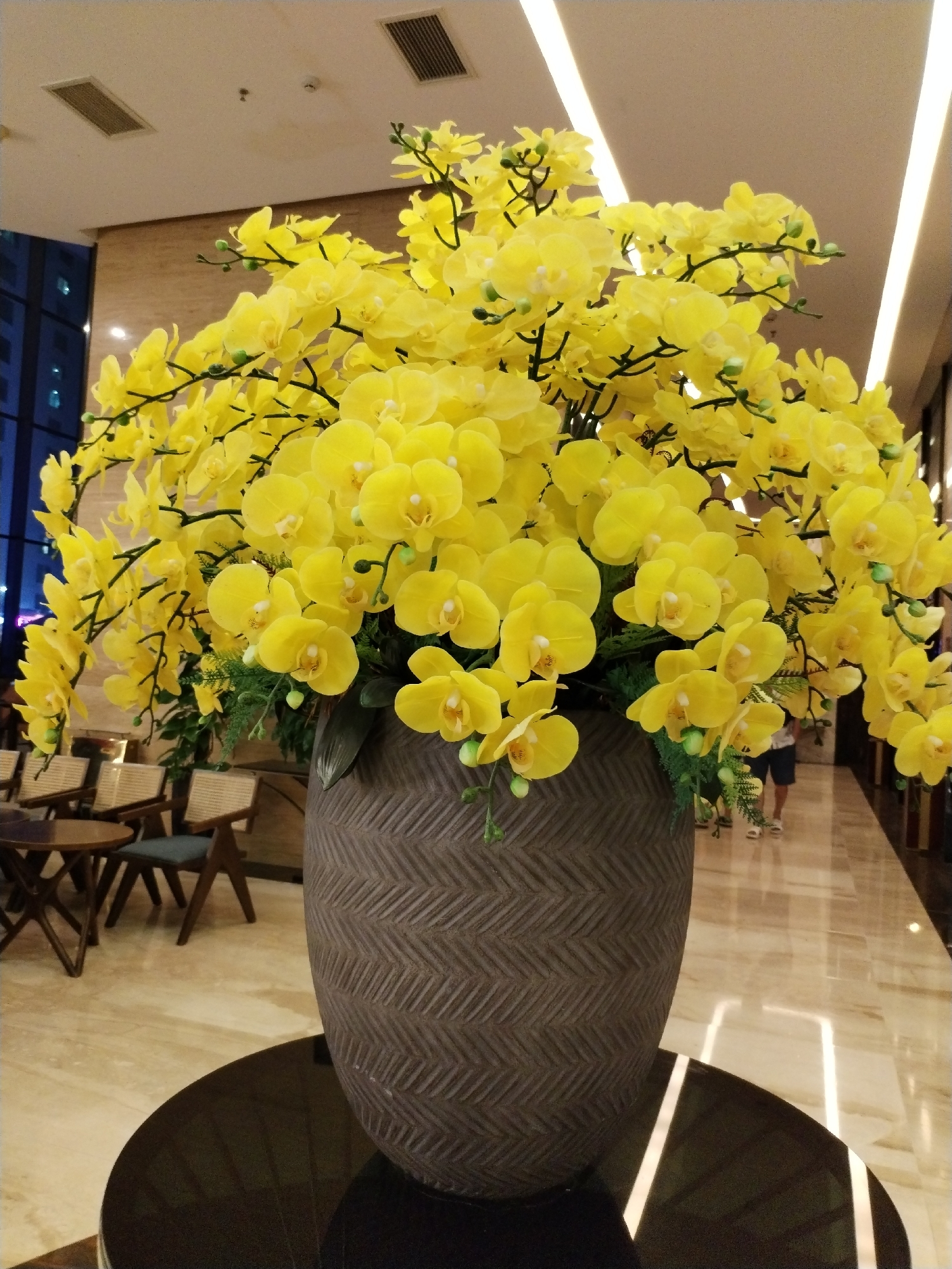 Captured indoors, the image showcases a large, ornate floral arrangement. Dominated by yellow orchids, the flowers are densely packed and spread generously from a sizable, textured brown vase. The vase sits on a glossy, dark-colored table, and a polished marble floor stretches behind it. To the left, there is a seating area with tables and chairs visible. In the background, a glimpse of the exterior reveals a building with visible windows, possibly indicating an urban setting. The lighting is bright, with ceiling lights reflected in the shiny floor. In the far background, indistinct figures are walking. Considering the presence of seating areas, building architecture, and interior design, the location suggests a hotel or upscale lobby. Based on the location description provided, it is implied that this image was taken in a hotel or public space in Bac Nha Trang Ward, Vietnam.
